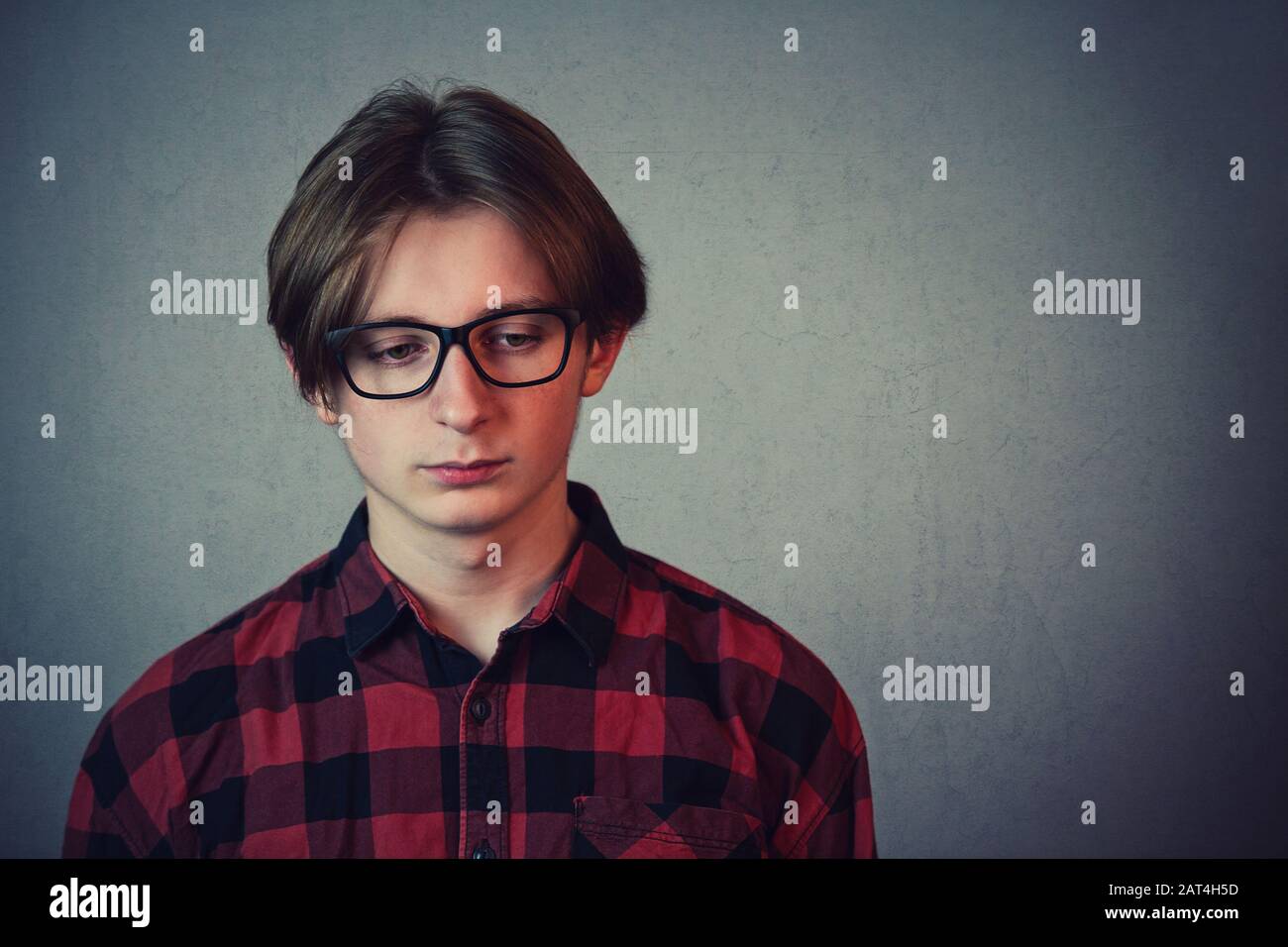 Triste e deluso ragazzo adolescente, indossando camicia rossa e occhiali, guardando verso il basso dispiaciuto isolato su sfondo grigio parete. Foto Stock