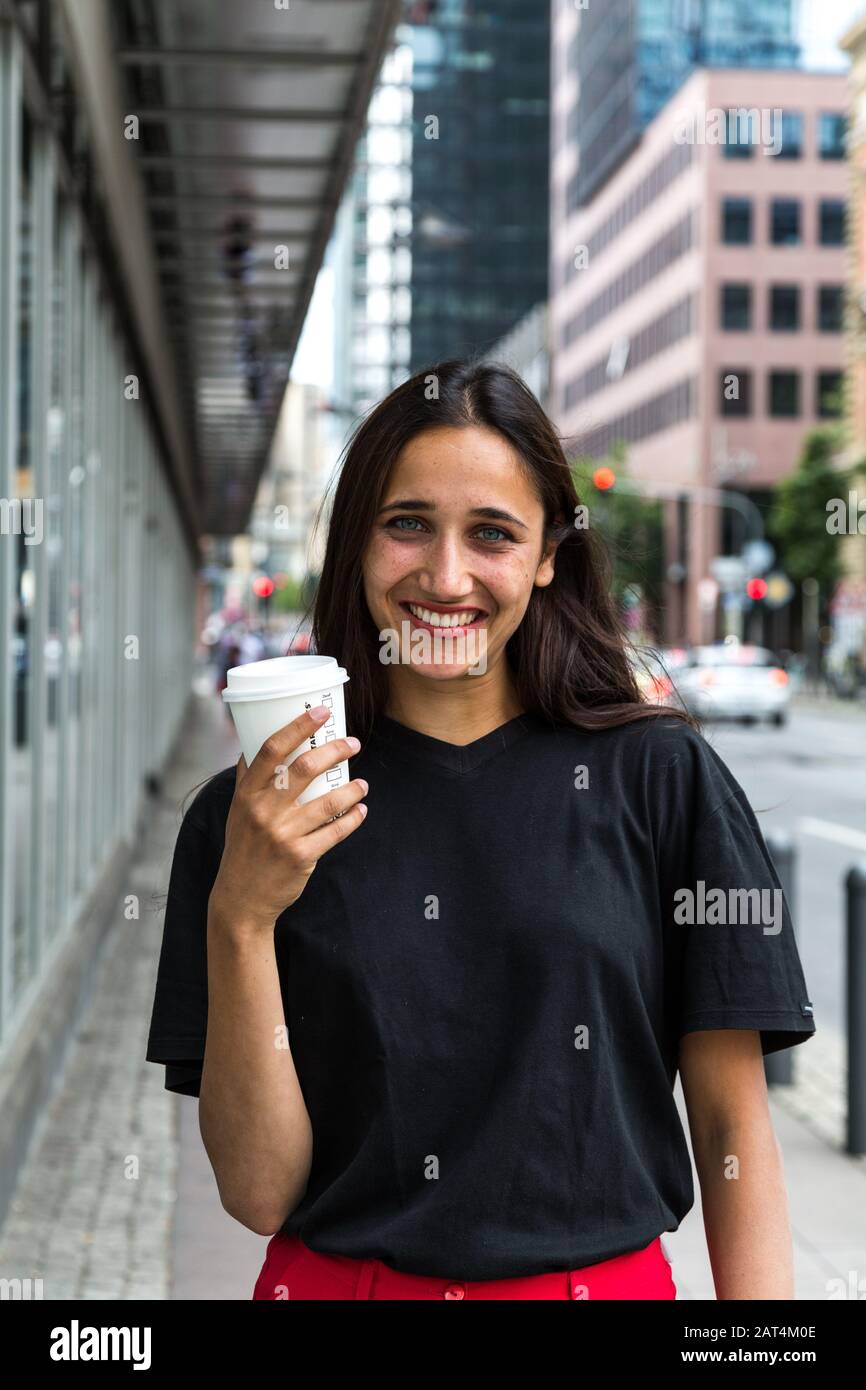 Ritratto di giovane donna sorridente ampiamente con tazza di carta in mano. Sfondo urbano. Scatto medio. Foto Stock