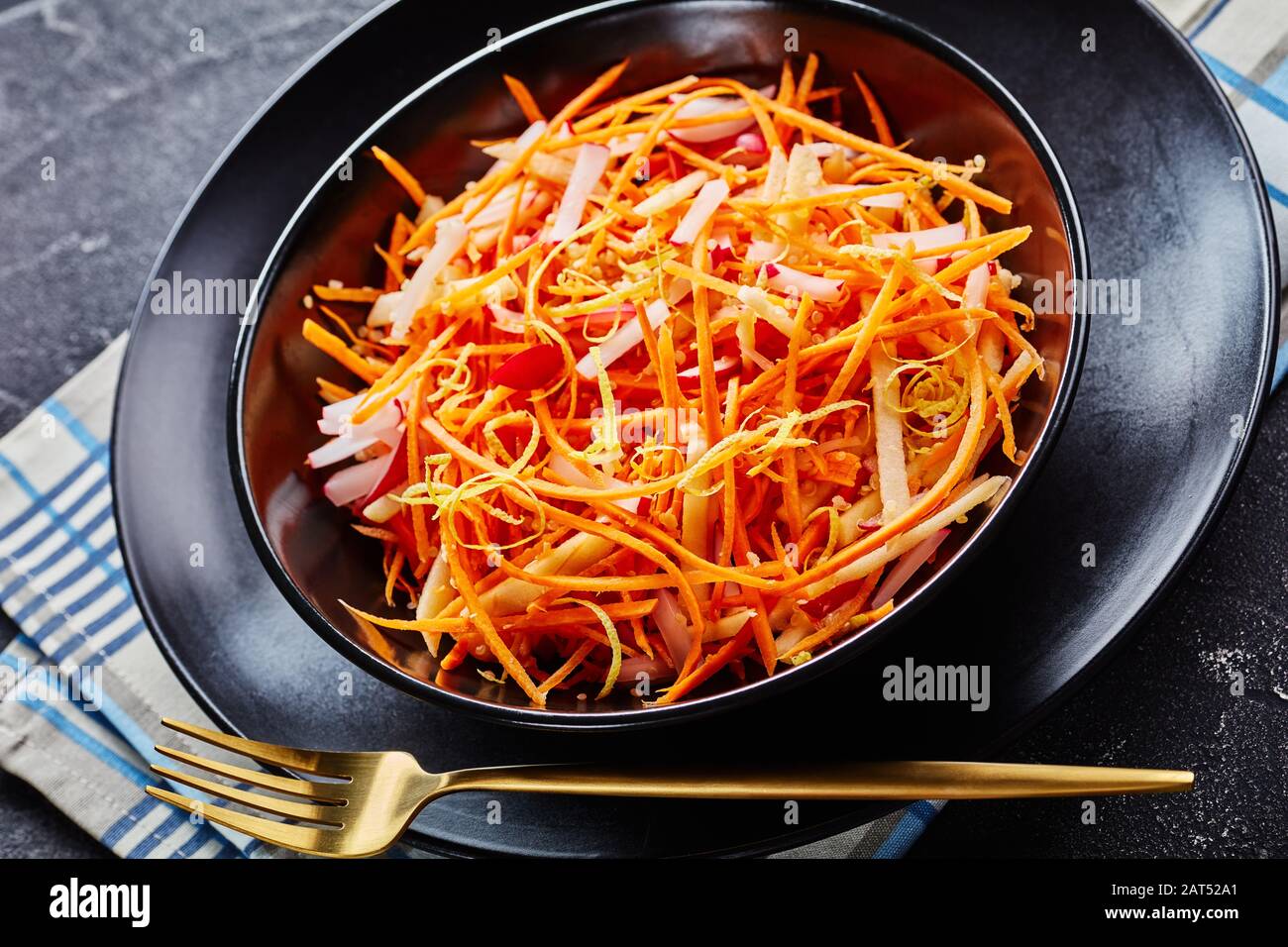 Primo piano di ravanello di carote Fresche e insalata di mele cosparse di semi di quinoa e scorza di limone in una ciotola nera con una forchetta dorata su un tavolo di legno, oriz Foto Stock