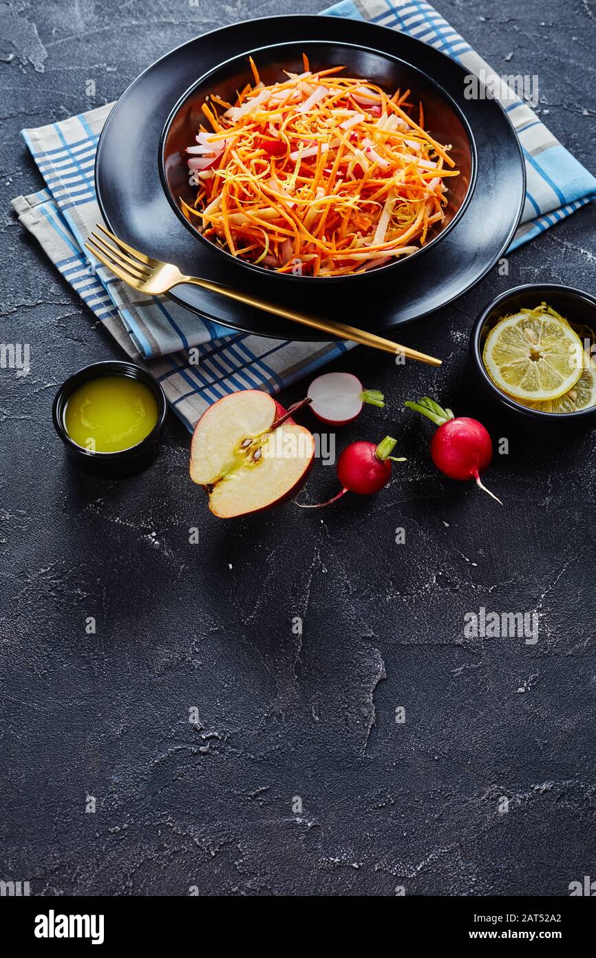 Primo piano di ravanello di carota Fresco e insalata di mele cosparsa di semi di quinoa e scorza di limone in una ciotola nera su un tavolo di legno con condimento fatto in casa, v Foto Stock