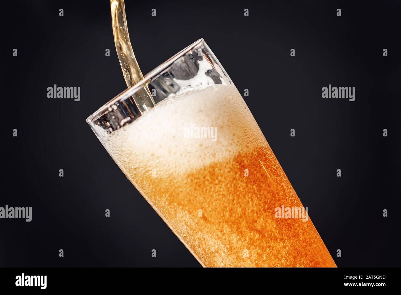 La birra fresca e leggera viene versata in un calice di vetro su sfondo scuro con spruzzi e schiuma Foto Stock