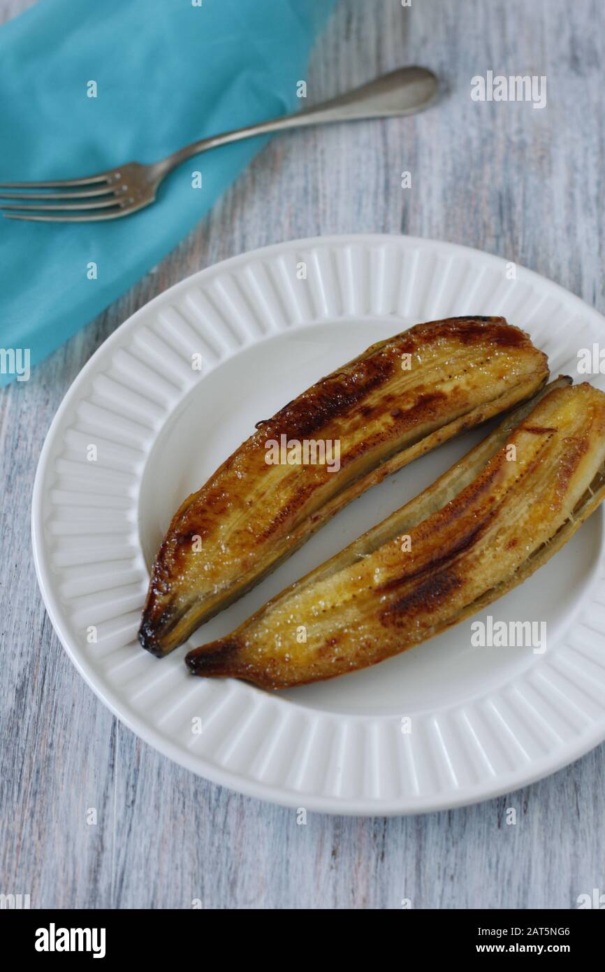 La banana tagliata a metà e cotta nella sua pelle fino al marrone dorato, servita su un piatto bianco. Scena con forchetta e tovagliolo blu Foto Stock