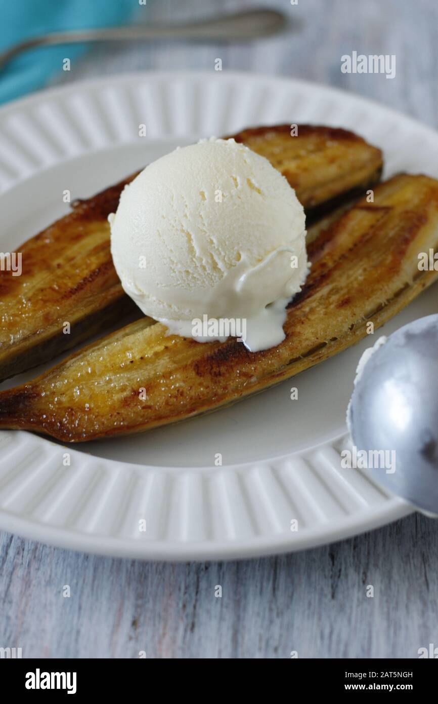 Banana cotta al forno dorato tagliata a metà con copertura di pelle con una paletta di gelato alla vaniglia su piastra bianca. Foto Stock