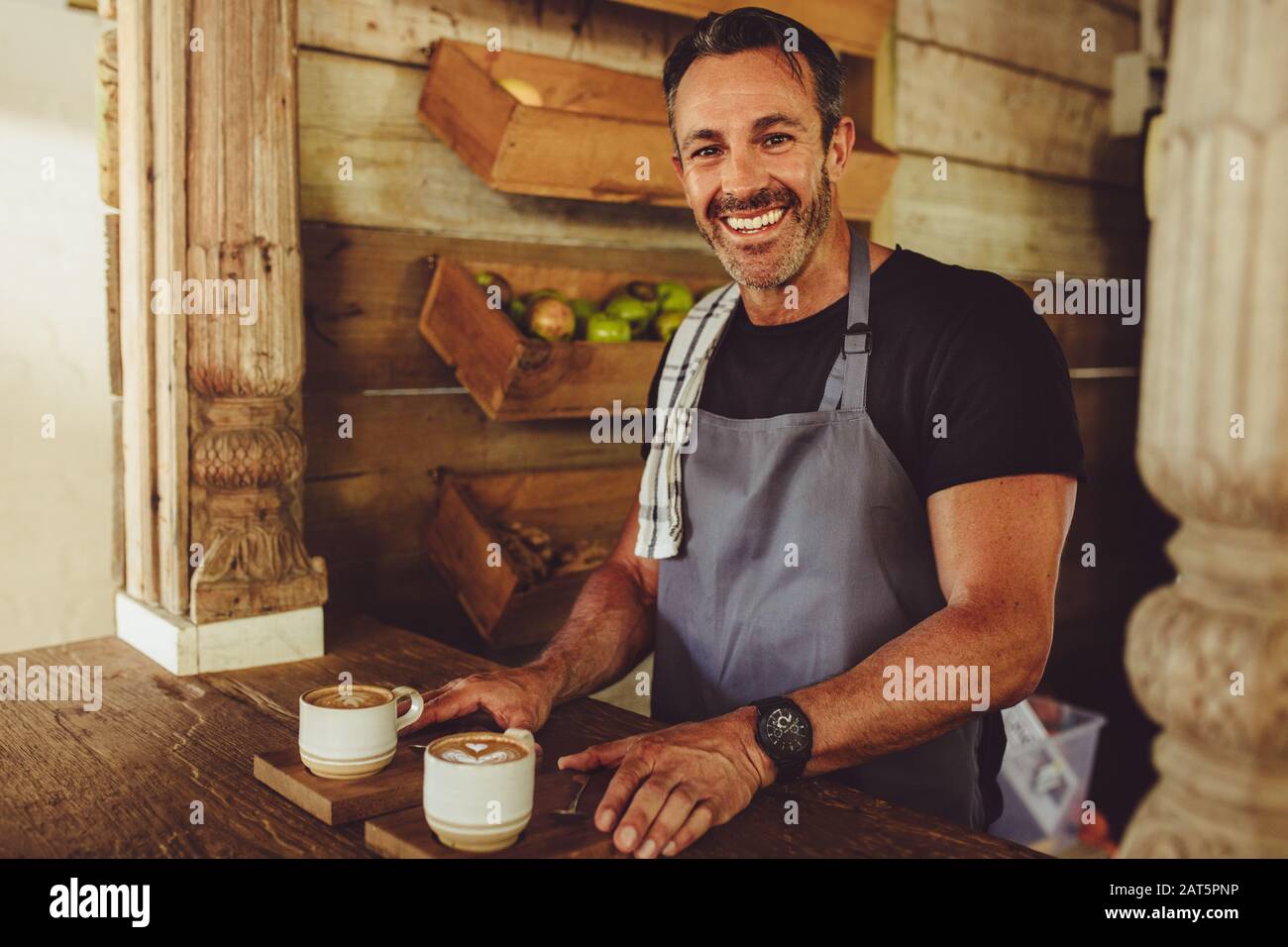 Ritratto di un barista maschile sorridente che serve caffè. Uomo che si trova dietro il bancone di un caffè che serve due tazze da caffè e sorridente. Foto Stock