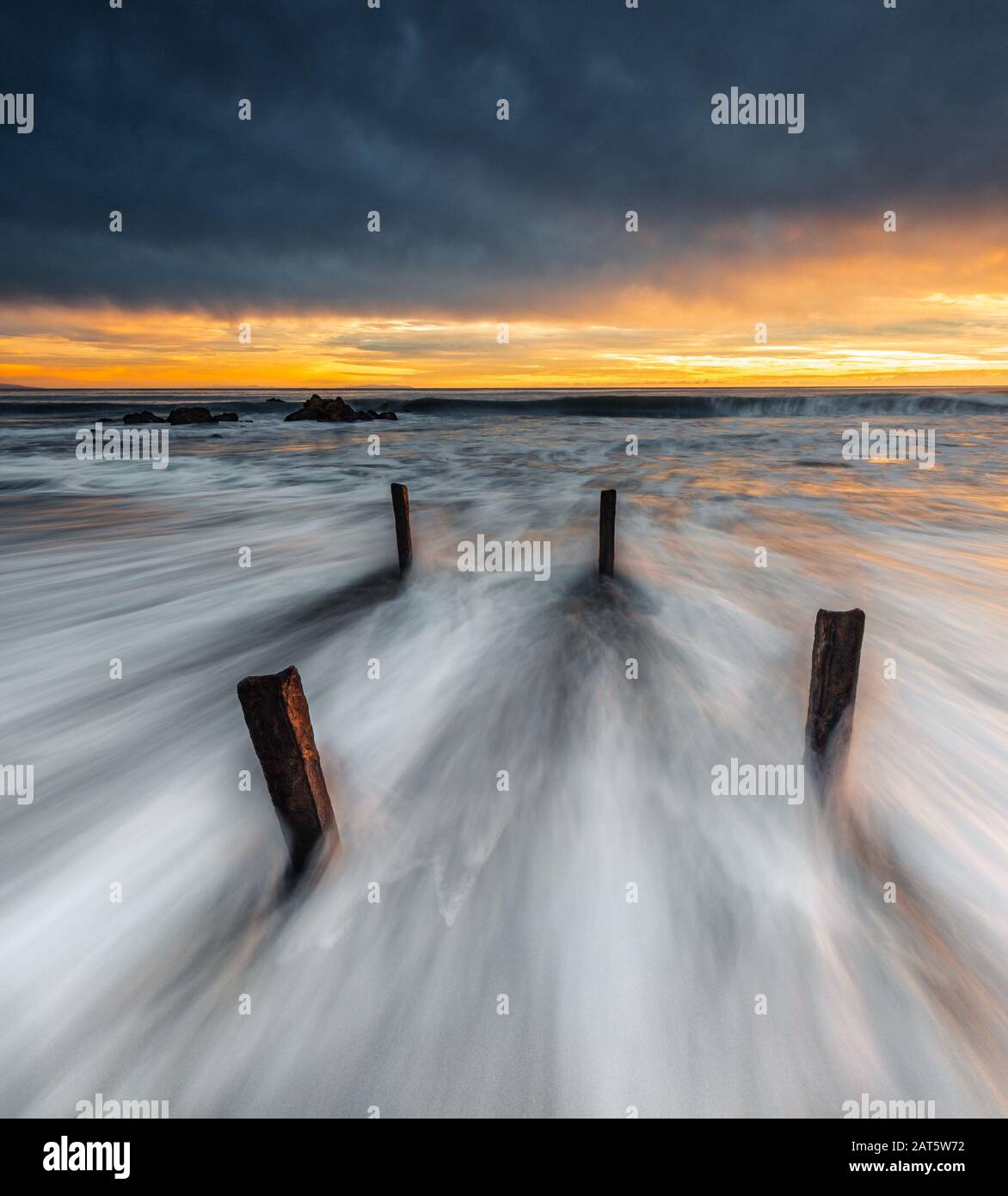 Con l'acqua bianca che si muove rapidamente oltre 4 piloni, l'immagine è bilanciata dai cieli pesanti pieni di pioggia sopra la striscia vibrante di luce dorata al tramonto. Foto Stock