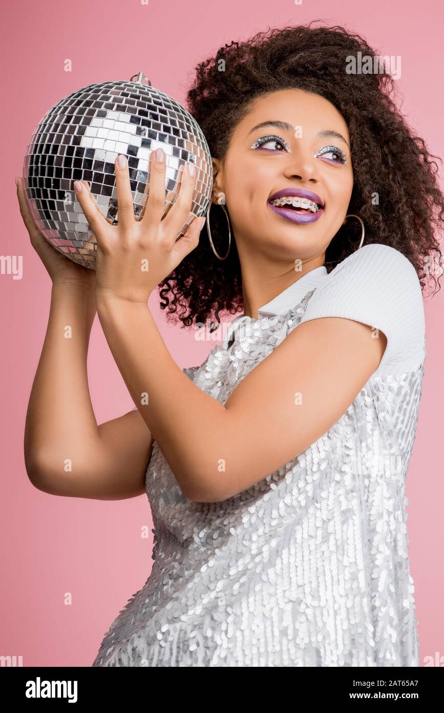 ragazza afroamericana sorridente in abito paillettes tenendo palla da discoteca, isolato su rosa Foto Stock