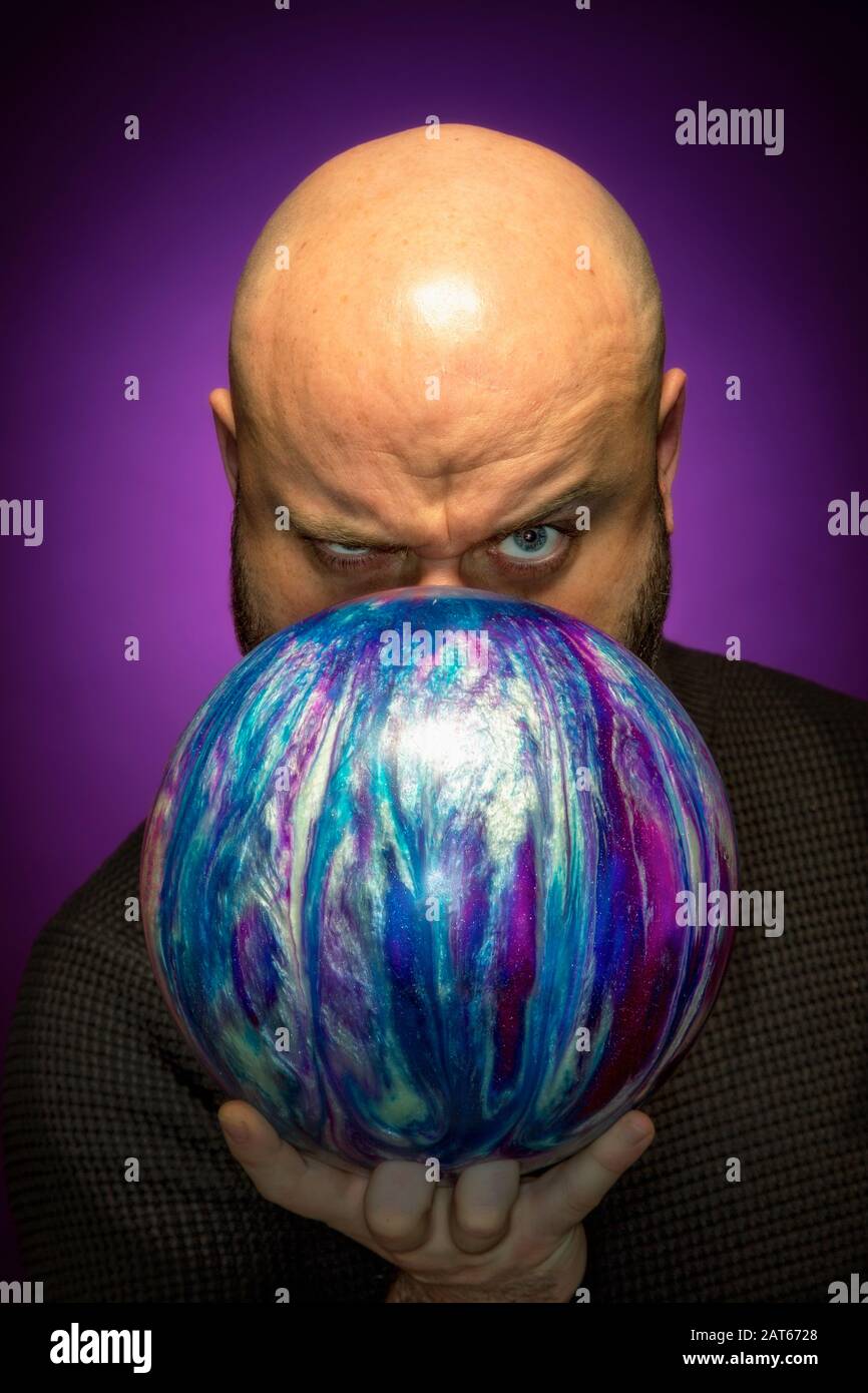 Un uomo calvo tiene una palla da bowling di fronte al suo volto schiacciando alla macchina fotografica come egli mira Foto Stock
