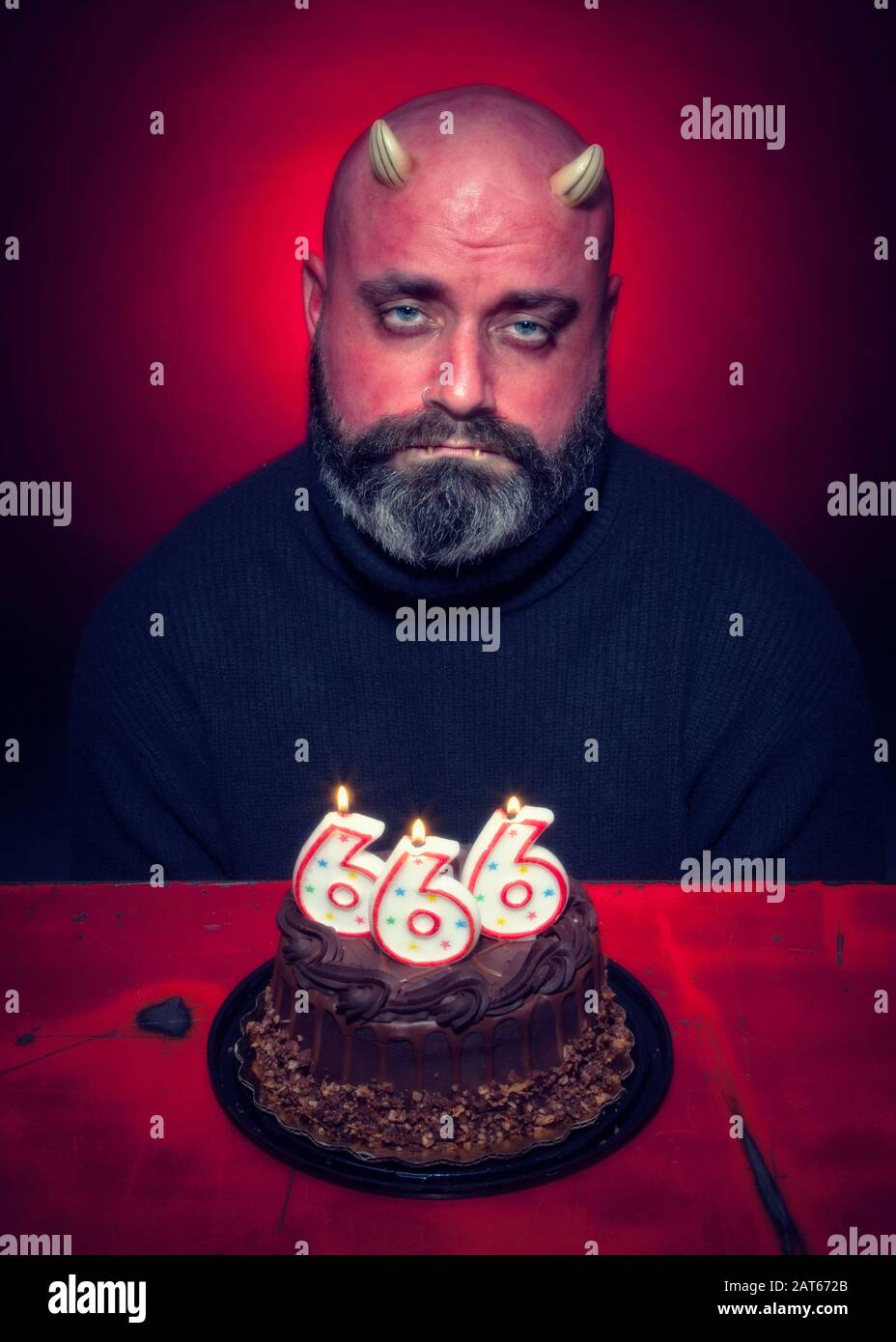 Un uomo triste siede davanti ad una torta di compleanno del cioccolato del diavolo con il numero 666 candele con uno sfondo rosso acceso Foto Stock