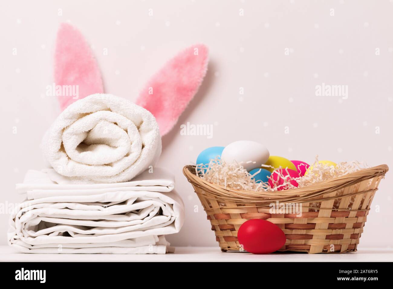 Un primo piano di una pila di lenzuola bianche pulite, asciugamani e orecchie di coniglio di Pasqua, un cesto di uova su un tavolo, su uno sfondo di pareti chiare. Foto Stock