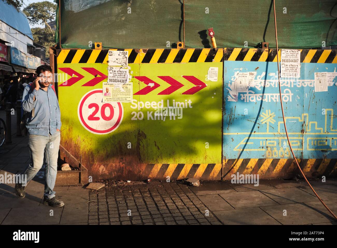 Al mattino presto, un pedone che utilizza il suo telefono cellulare passa il sito di costruzione di una nuova stazione della metropolitana Mumbai, Mumbai, India Foto Stock