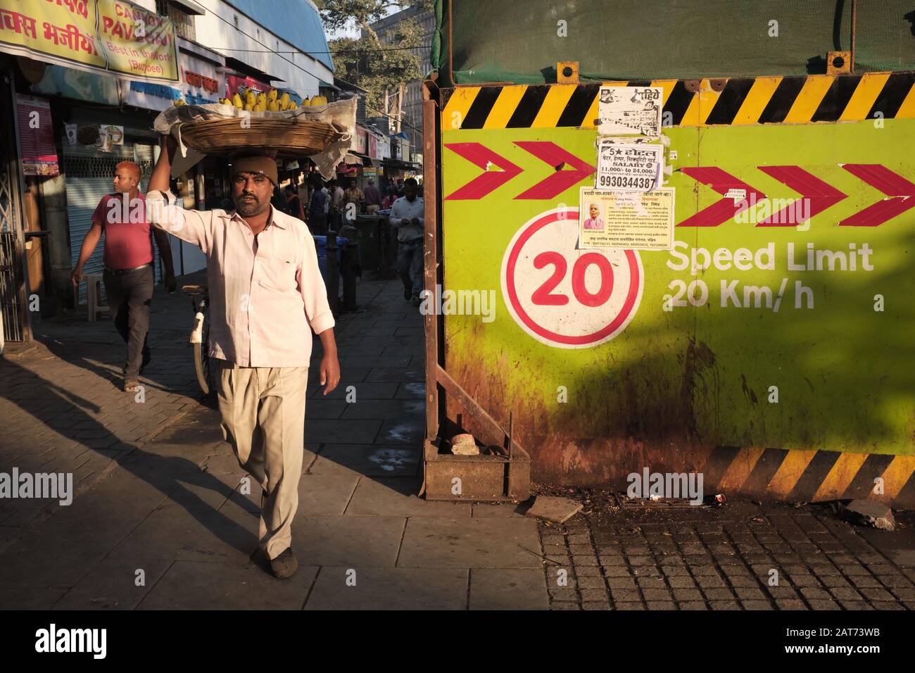 La mattina presto, un venditore di frutta che porta un cesto di banane sulla sua testa passa un cantiere di costruzione di una nuova stazione della metropolitana di Mumbai; Mumbai, India Foto Stock