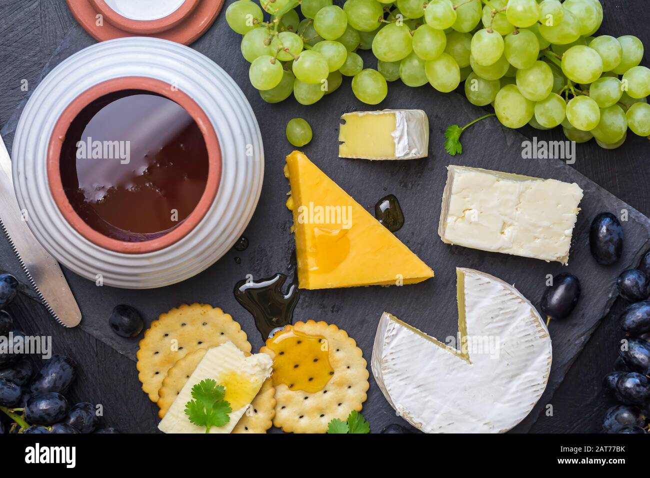 Tagliere di formaggio con formaggio assortito, camembert, cheddar, gouda, cracker, uva e vaso di miele su sfondo nero ardesia - foto in alto Foto Stock