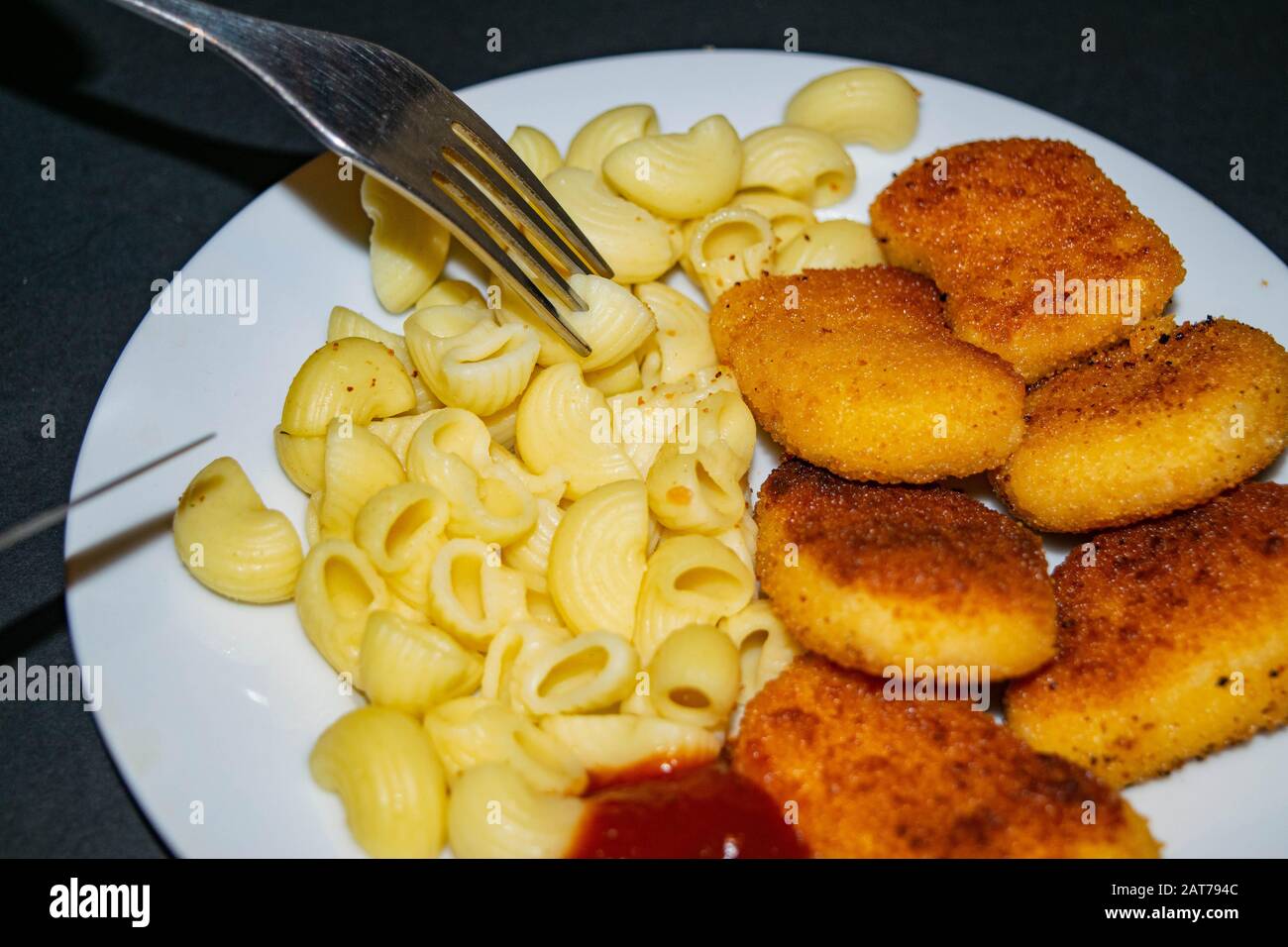 Pepite di pollo fritte, pasta bollita, ketchup e forchetta su un piatto bianco su uno sfondo scuro. Primo piano Foto Stock