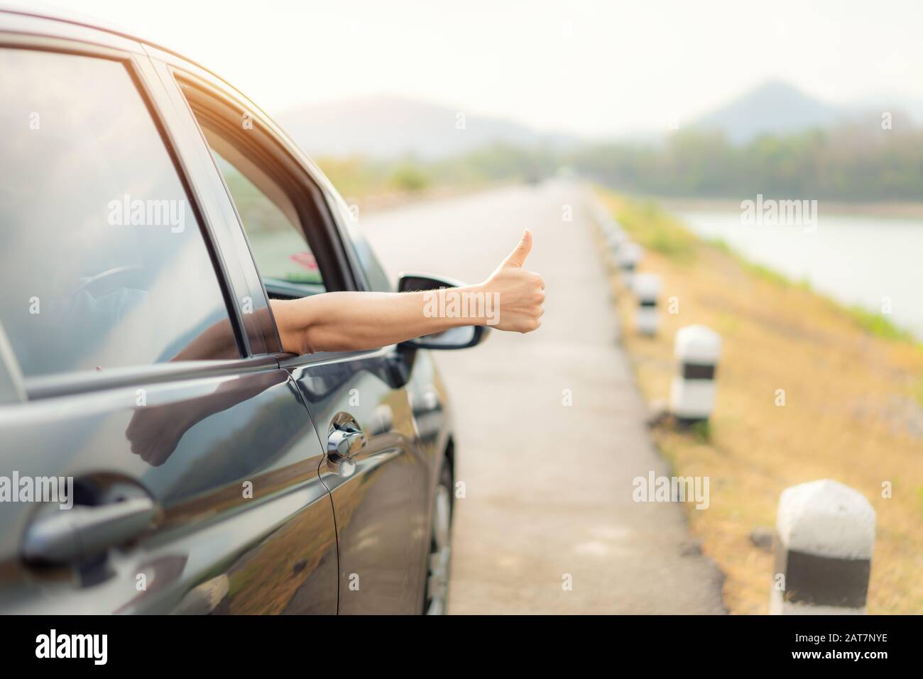 Mano di uomo viaggiatore con pollici in su dall'automobile forma la strada di asfalto va viaggiare con il lago di paesaggio bello e le montagne e la luce del sole in sera. C Foto Stock