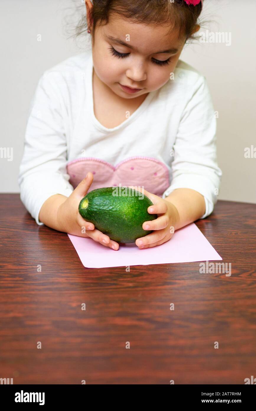 Piccola cute sorridente ragazza capelli neri tenendo avocado verde. Concetto di cibo sano per i bambini. Foto Stock