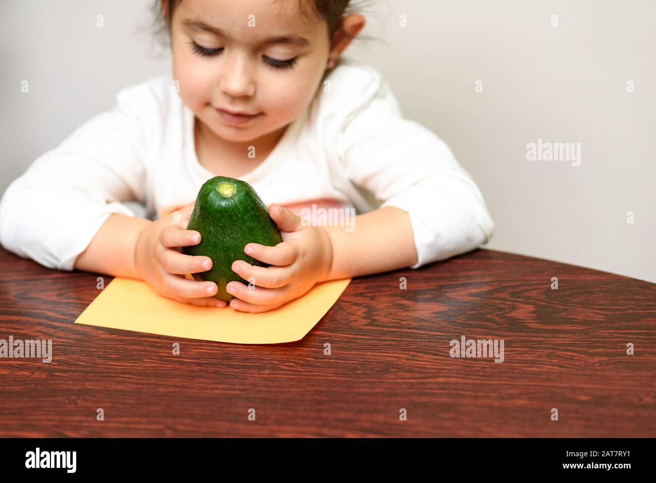 Piccola cute sorridente ragazza capelli neri tenendo avocado verde. Concetto di cibo sano per i bambini. Focus selettivo su avokado. Foto Stock