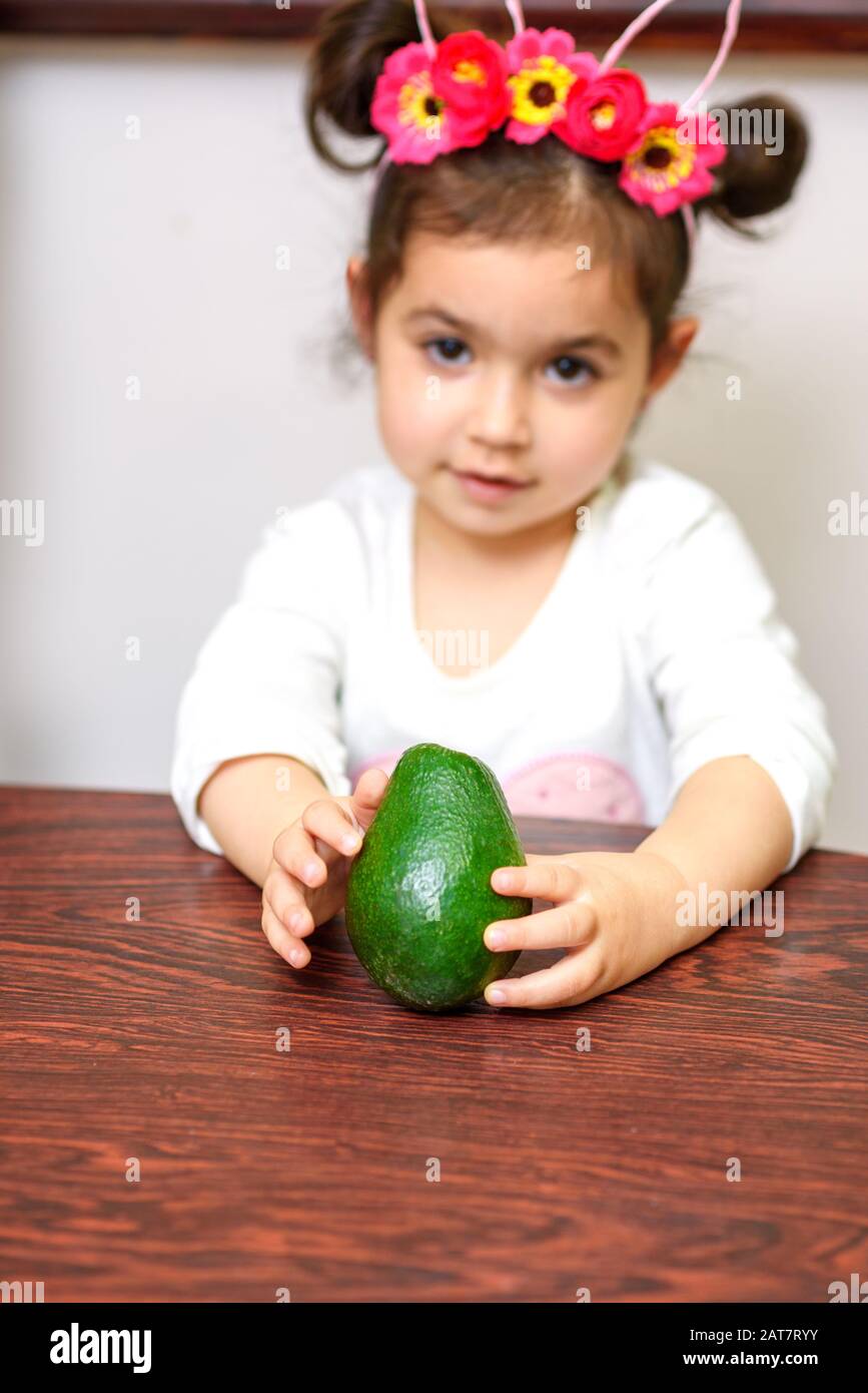 Piccola cute sorridente ragazza capelli neri tenendo avocado verde. Concetto di cibo sano per i bambini. Focus selettivo su avokado. Foto Stock