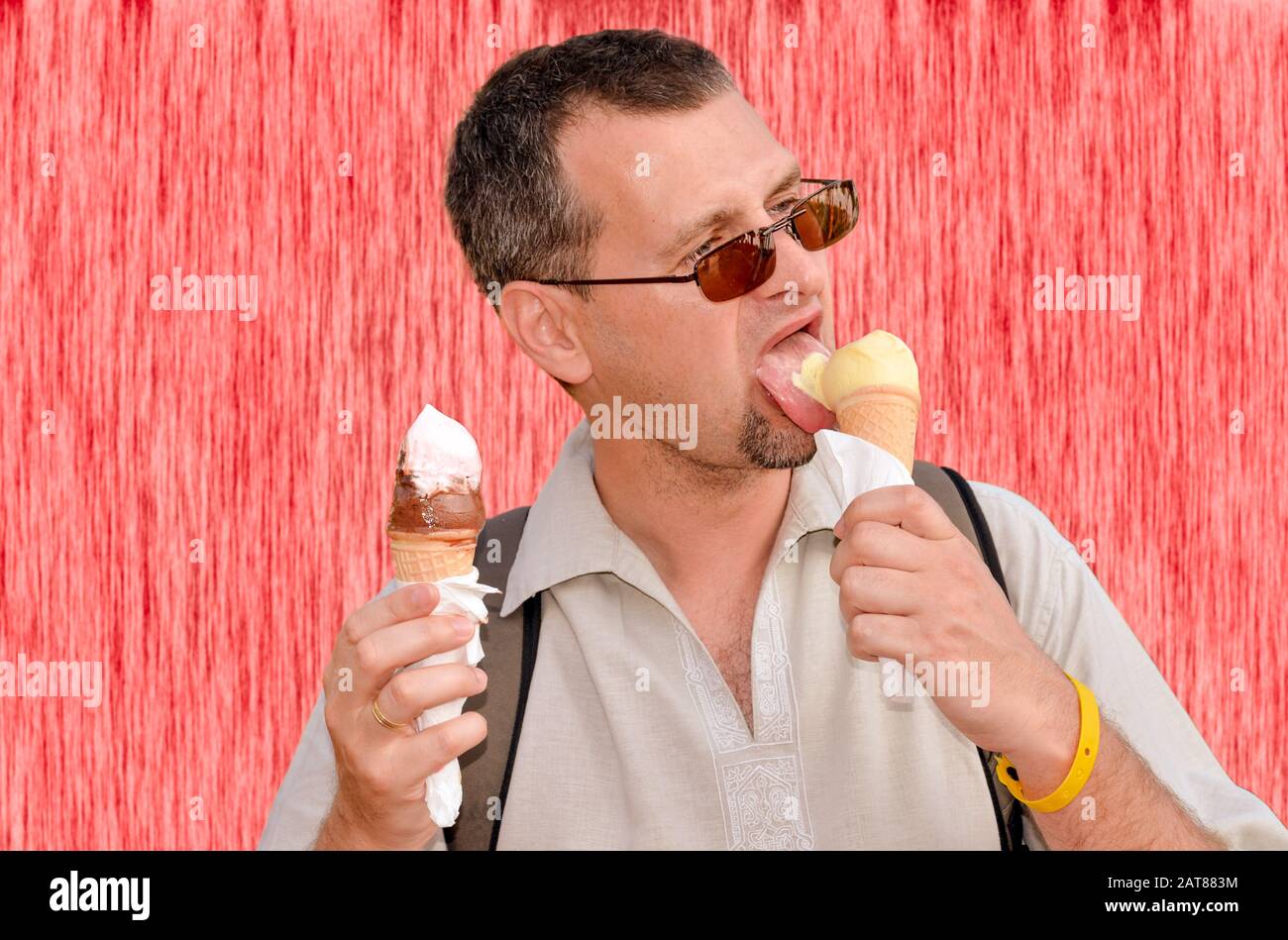 Ritratto di un uomo che mangia due gelati in coni di wafer in entrambe le mani sulla parete rossa di fondo Foto Stock