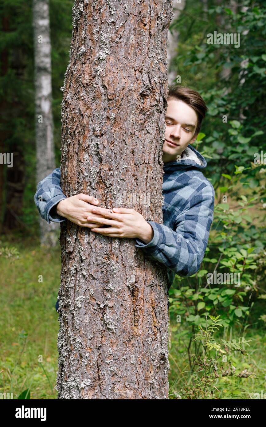 Ragazzo adolescente che abbraccia l'albero di pino in una foresta estiva appoggiato al tronco dell'albero con occhi chiusi che abbracciano la natura Foto Stock