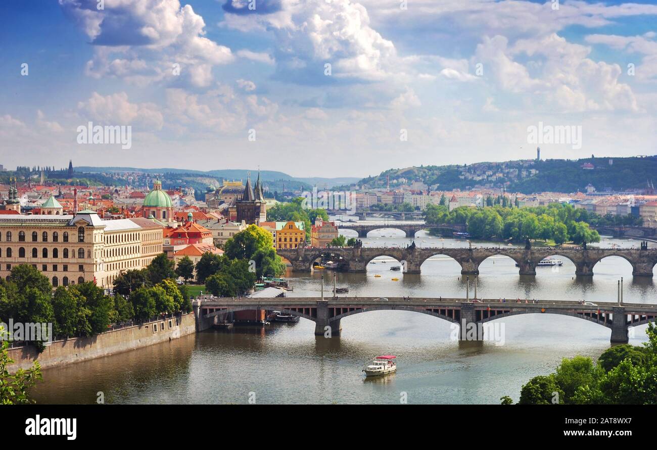 Vista del Ponte Carlo e altri ponti ad arco, alberi verdi con molte piccole case sotto tetti rossi vicino al fiume Moldava contro il vibrante cielo blu e whi Foto Stock