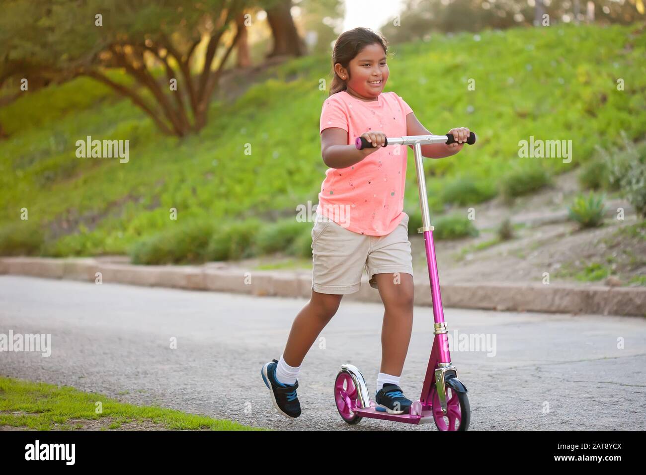 Una bambina che guida uno scooter rosa con fiducia in una strada fiancheggiata da alberi e erba. Foto Stock