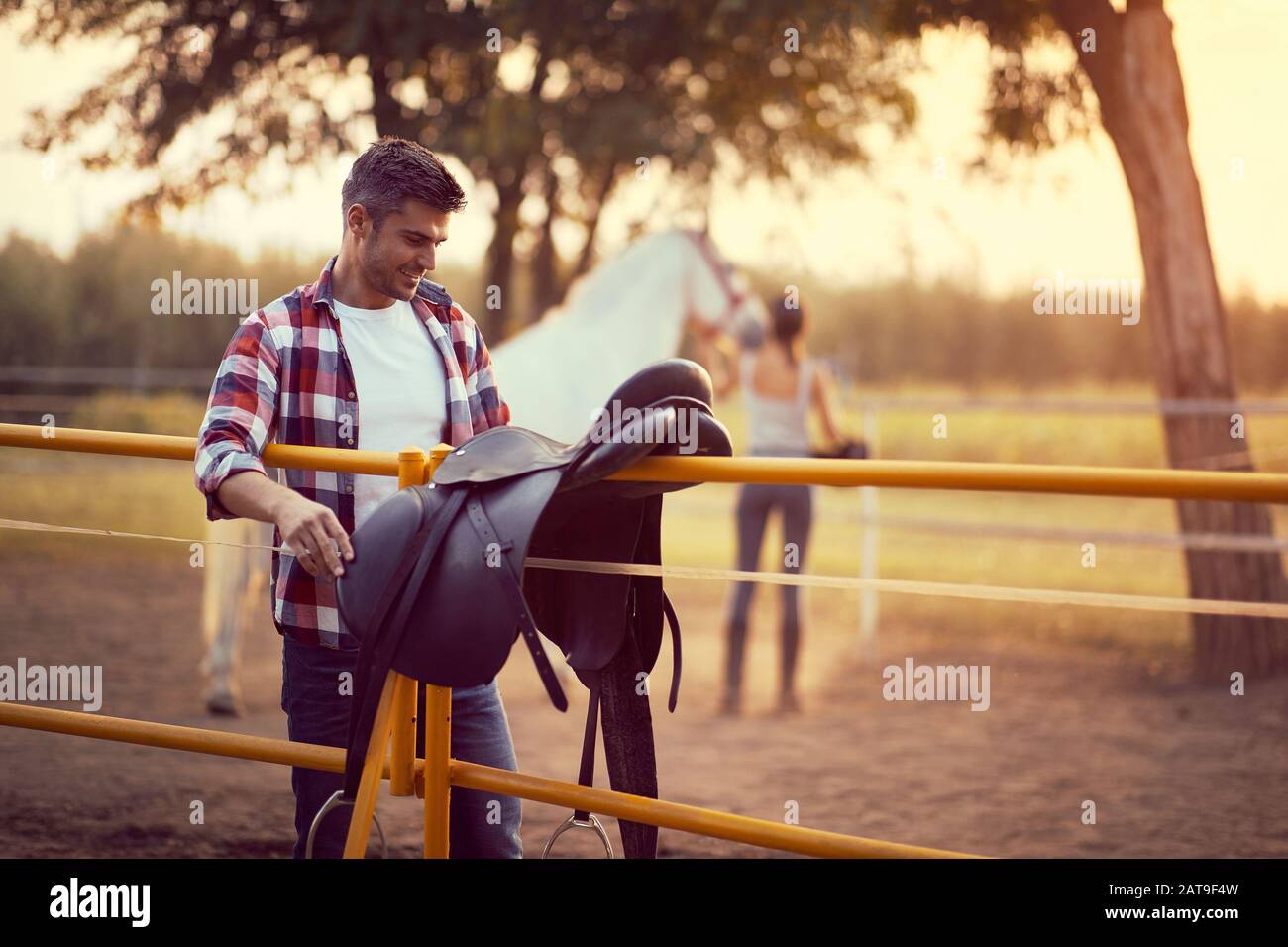 Uomo che prepara una sella di cavallo per un giro, donna con un cavallo nella parte posteriore. Allenamento in campagna, ora d'oro al tramonto. Libertà natura concetto. Foto Stock