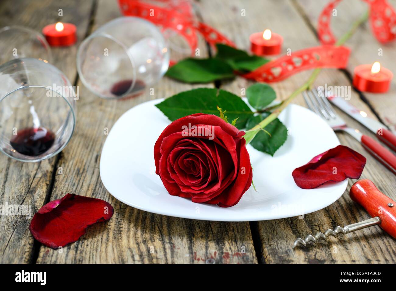 La rosa sul piatto, due bicchieri di vino, cavatappi, coltello, forchetta e candele brucianti su un vecchio tavolo di legno. Messa a fuoco selettiva. Foto Stock