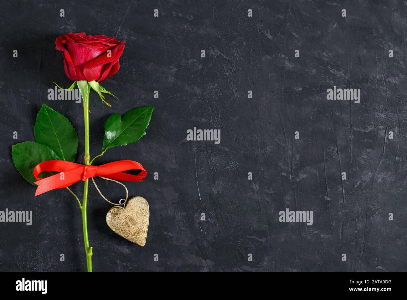 Romantica rosa rossa, cuore in legno e nastro rosso su lavagna nera scura con spazio per il testo. Concetto di carta di giorno di San Valentino. Foto Stock