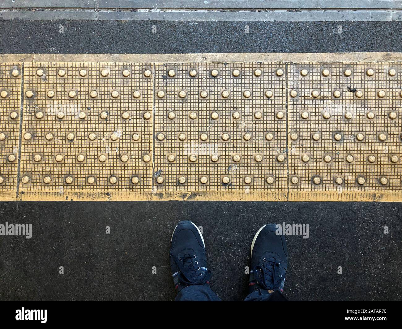 Attenzione al divario: In attesa sulla piattaforma di una stazione della metropolitana di Londra. POV, guardando giù la linea di divisione goffrata, il pavimento e le scarpe Foto Stock