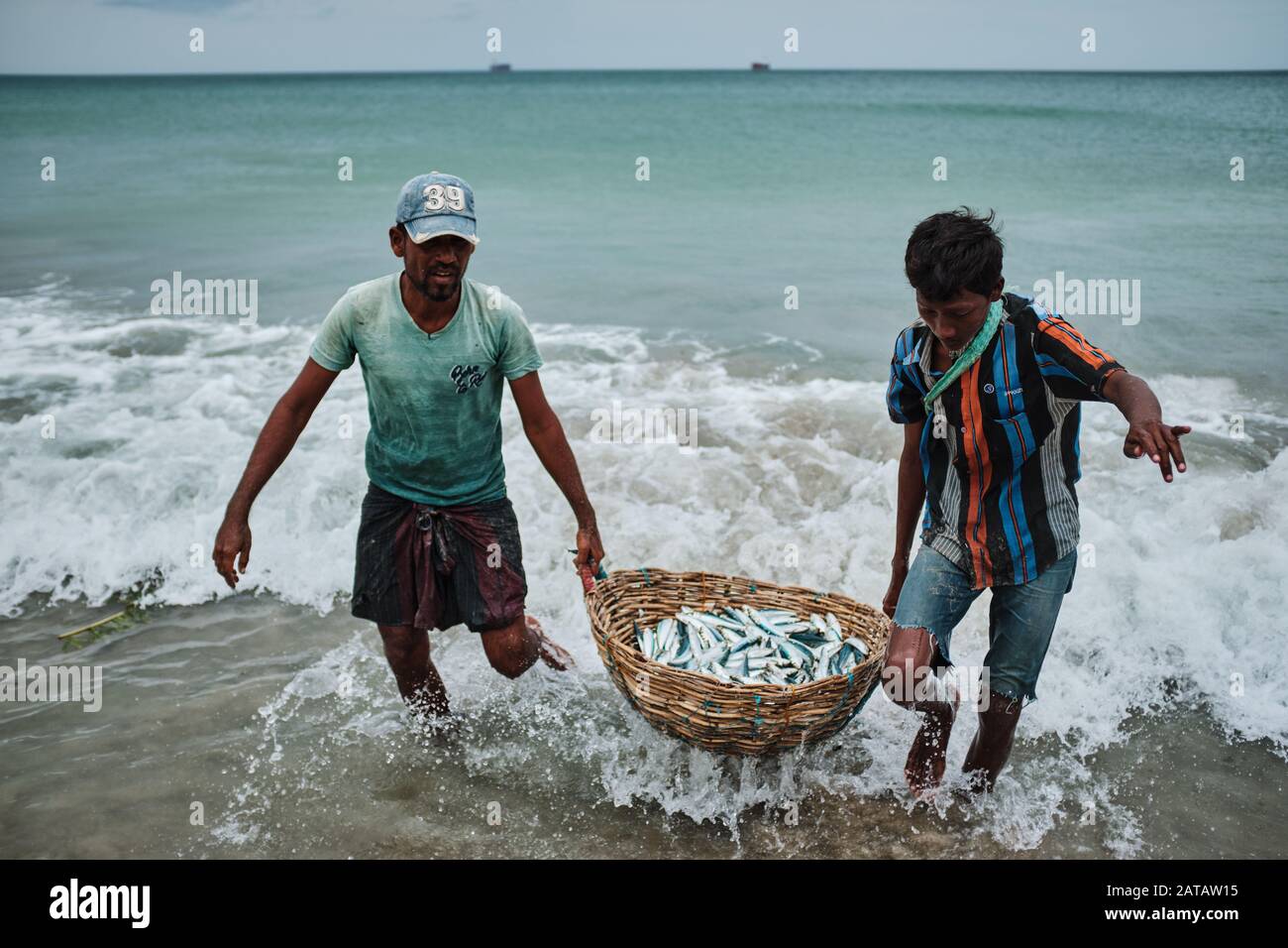 Due uomini dello Sri Lanka che trasportano il pesce in un cestino di bambo sulla spiaggia di Trincomalee e lavarlo nell'oceano. Foto Stock