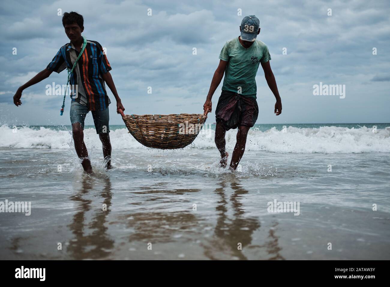 Due uomini dello Sri Lanka che trasportano il pesce in un cestino di bambo sulla spiaggia di Trincomalee e lavarlo nell'oceano. Foto Stock