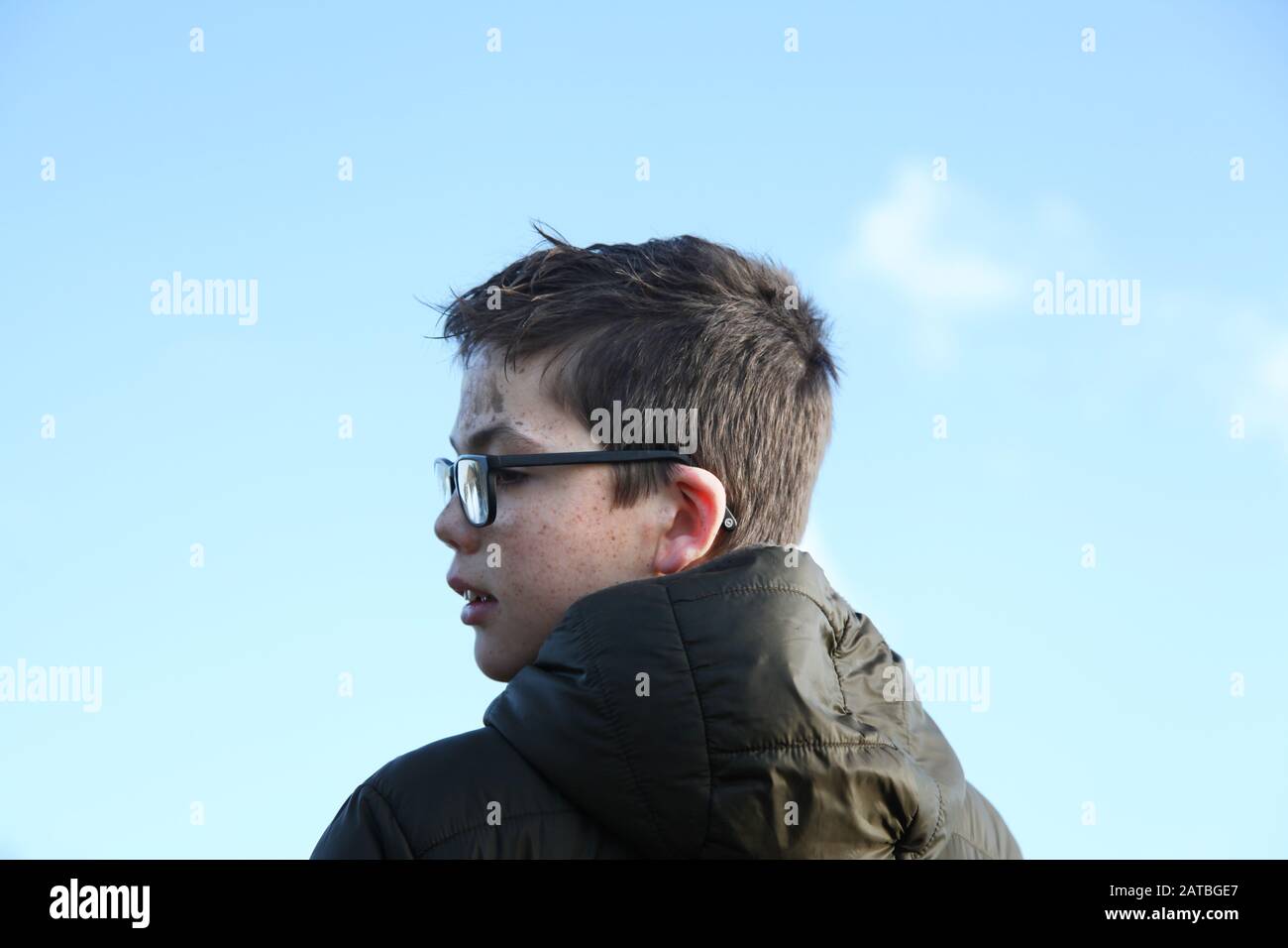 Un giovane ragazzo di 12 in occhiali con un viso fangoso che guarda al lato con sfondo blu cielo Foto Stock