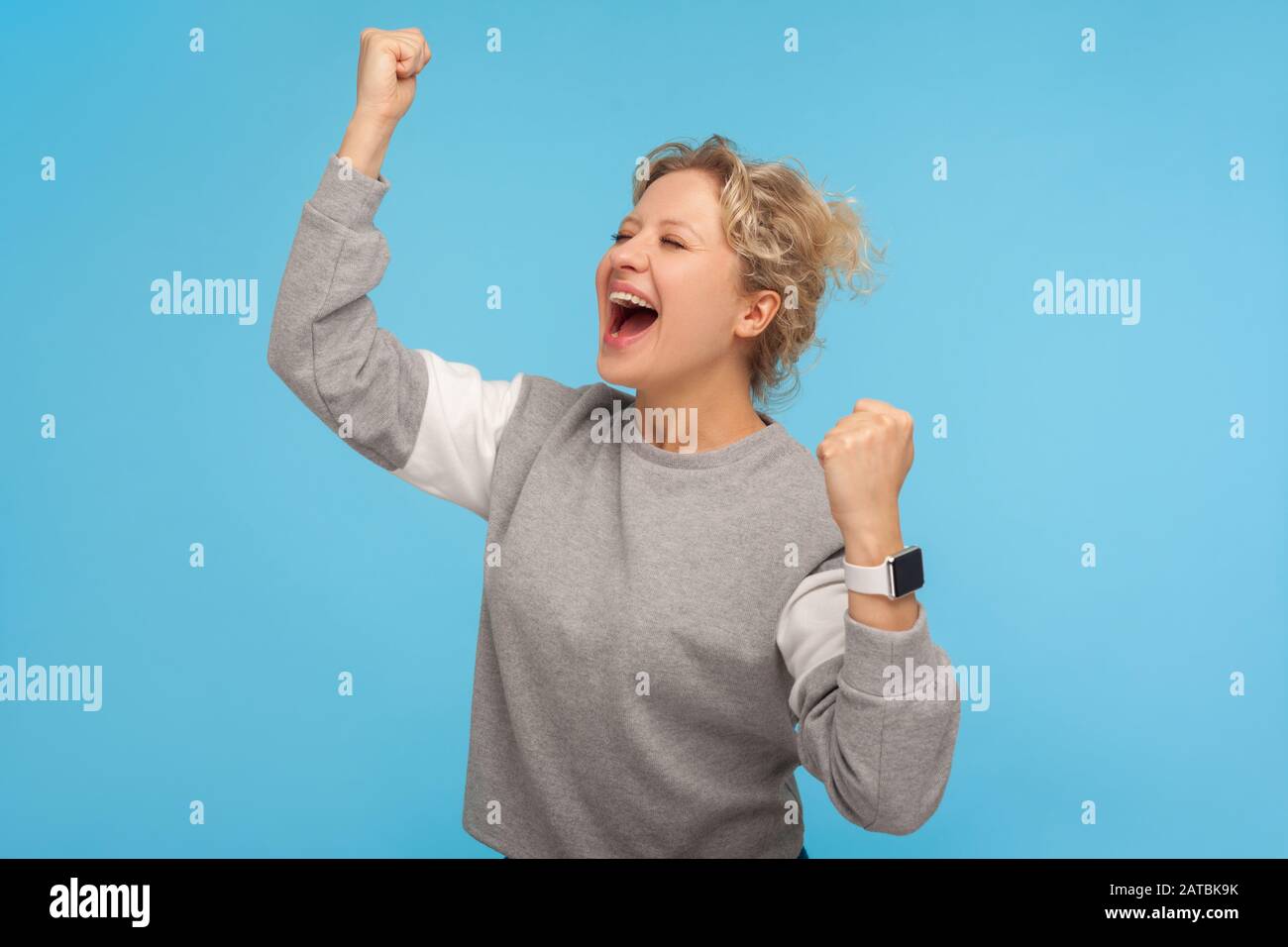 Entusiasta estatica donna con capelli corti ricci in felpa gridando sì e alzando le mani, esprimendo molta gioia di vincere, celebrando il successo. Io Foto Stock