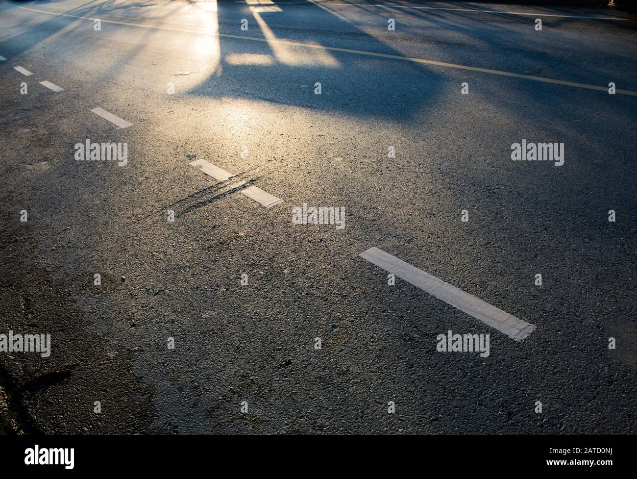 La luce del mattino cade su una superficie di asfalto vuota, con ombre, segnaletica stradale bianca e gialla e irregolarità di superficie di interesse. Foto Stock