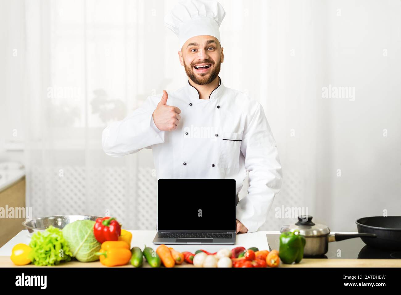 Chef Che Mostra Notebook Schermo Vuoto Gesturante Thumbs-Up Standing In Cucina Foto Stock
