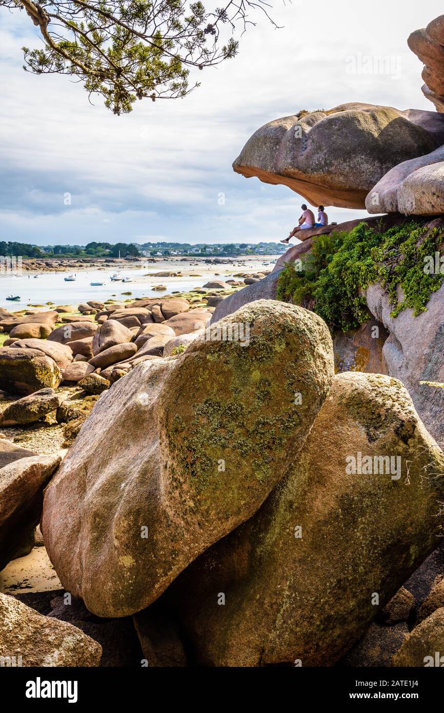 Due giovani uomini seduti sotto un masso di granito rosa equilibrato godono la vista sulla baia di Sainte-Anne a bassa marea in Tregastel, Bretagna, Francia. Foto Stock