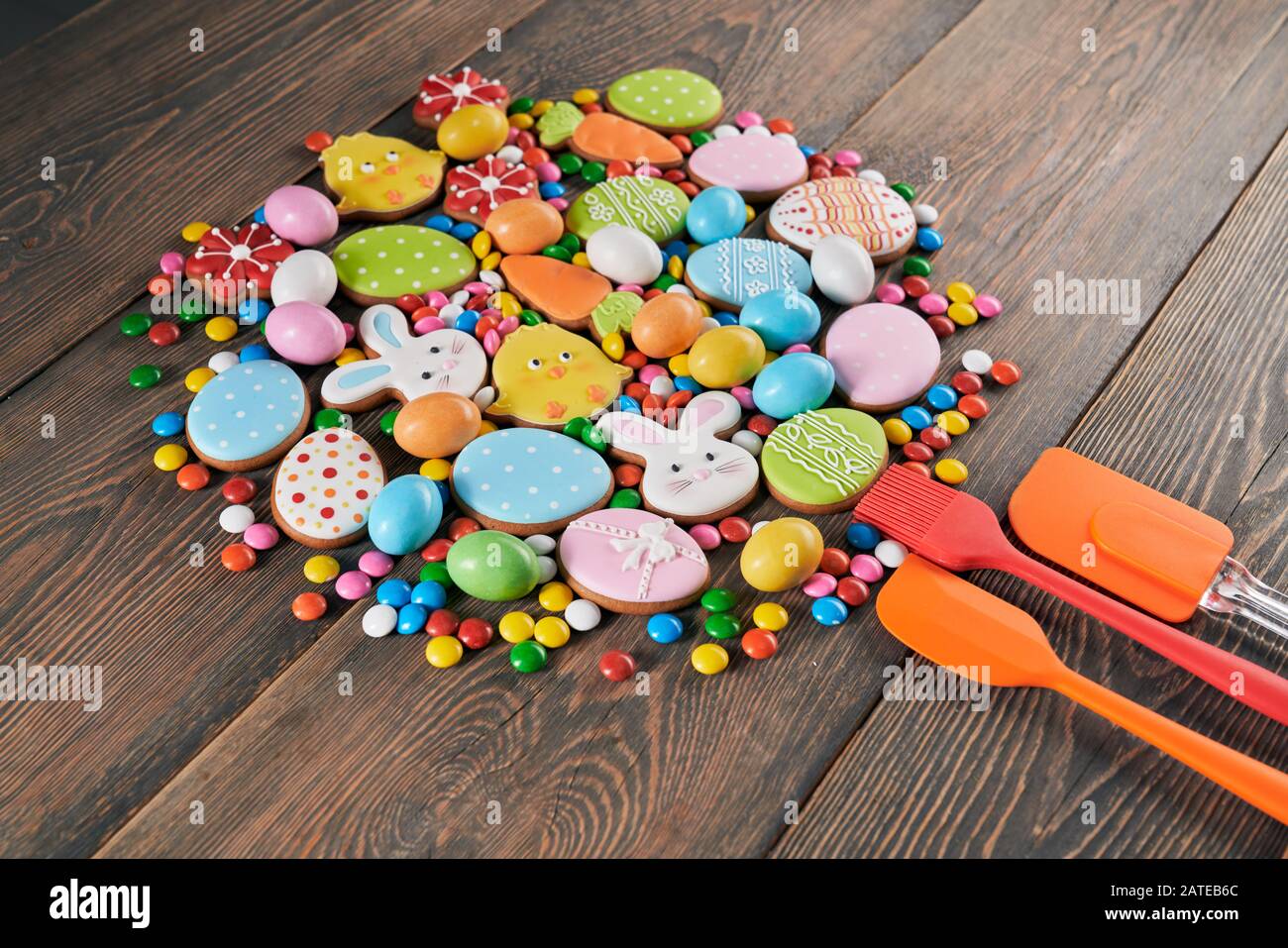 Biscotti colorati glassati allo zenzero e palline di cioccolato con guscio di zucchero isolato su tavolo di legno. Dolce fatto in casa a forma di animali pasquali, uova e carote, tre utensili da cucina in silicone nelle vicinanze. Foto Stock