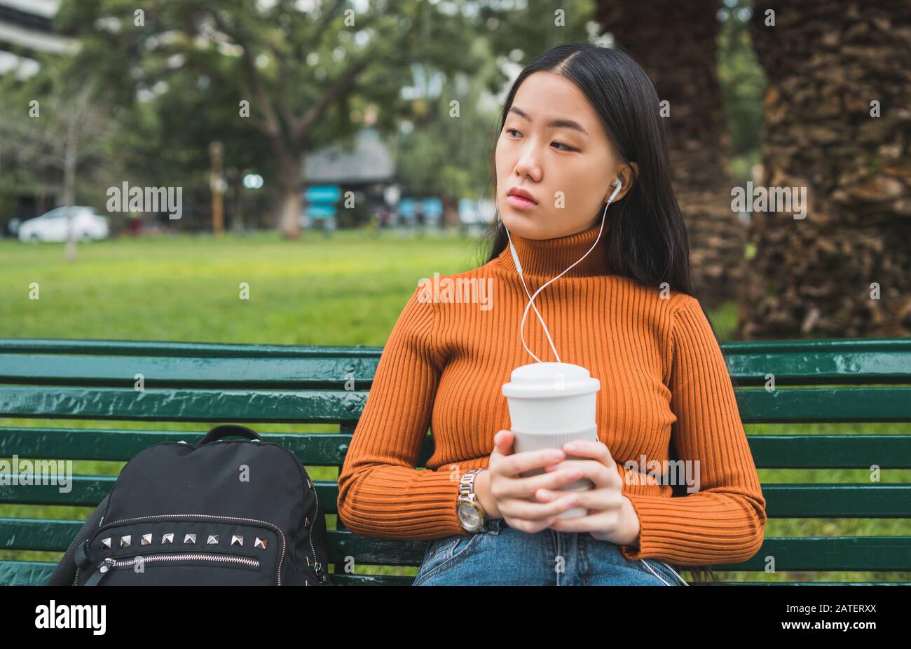 Ritratto di giovane donna asiatica ascoltando musica con auricolari e tenendo una tazza di caffè nel parco all'aperto. Foto Stock