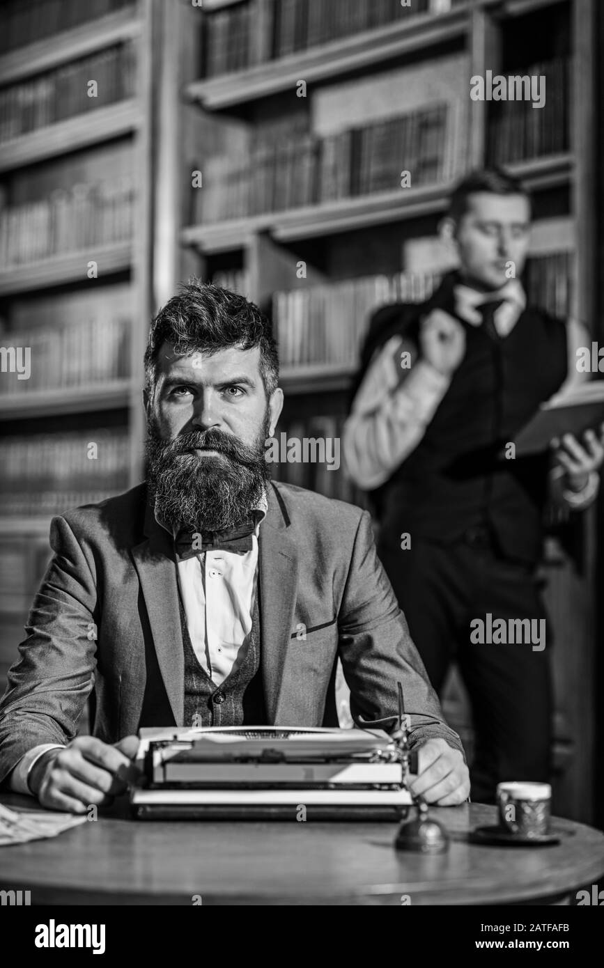 Scrittrice bearded con tipizzazione del volto concentrata su una macchina da scrivere di fronte alla biblioteca, vicino al suo amico, poeta o ricercatore. Scrittore lavora con macchina da scrivere, defocused Foto Stock