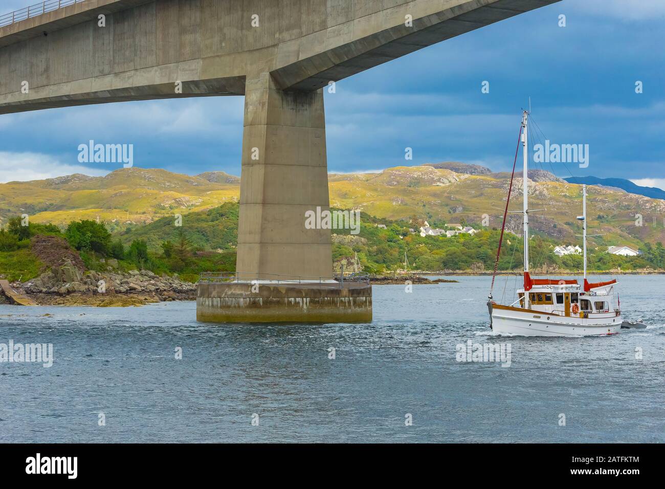Ponte dell'Isola di Skye con imbarcazione da crociera bianca e rossa che passa sotto il famoso ponte. Montagne e cottage sullo sfondo. Orizzontale. Foto Stock