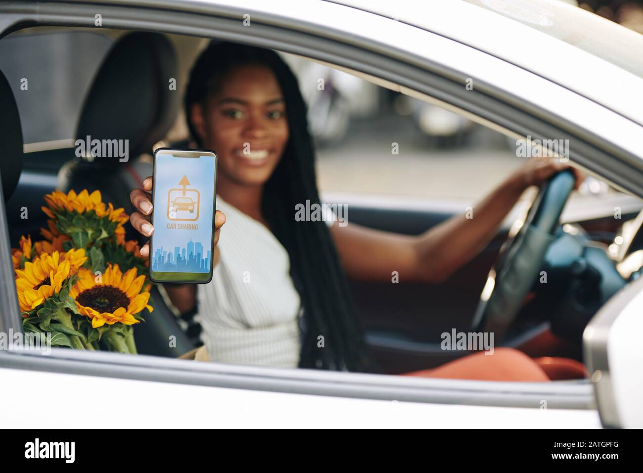 Giovane donna nera sorridente, che guida l'auto e mostra lo smartphone con l'applicazione di condivisione dell'auto Foto Stock