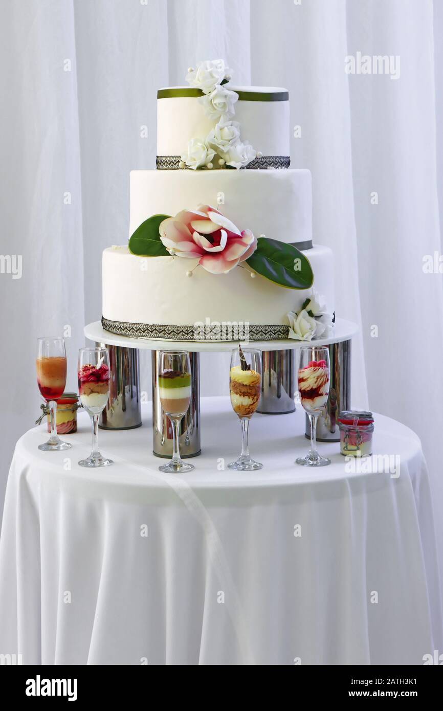 Grande torta nuziale con tre piani decorata da rose bianche e rosa. Alcuni bicchieri con dessert davanti alla torta. Foto Stock