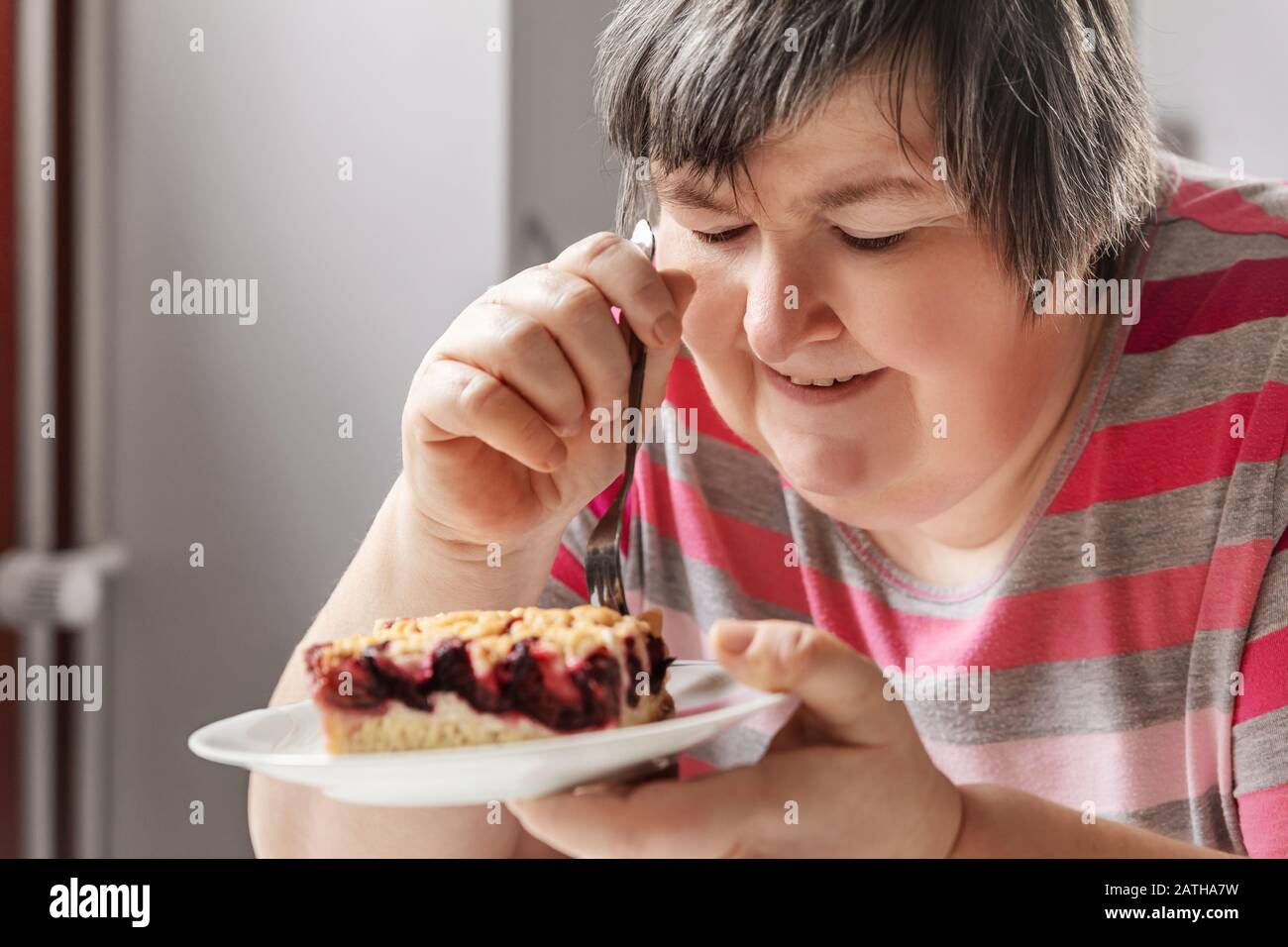 la donna con disabilità mentale sta mangiando una torta di prugne mentre si siede in cucina Foto Stock