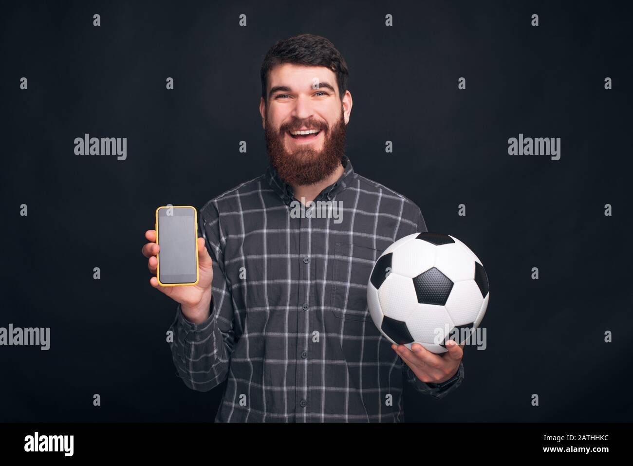 Puoi guardare il calcio o il calcio che si stringe sul tuo telefono. L'uomo bearded sta tenendo una sfera ed un telefono nell'altra mano. Foto Stock