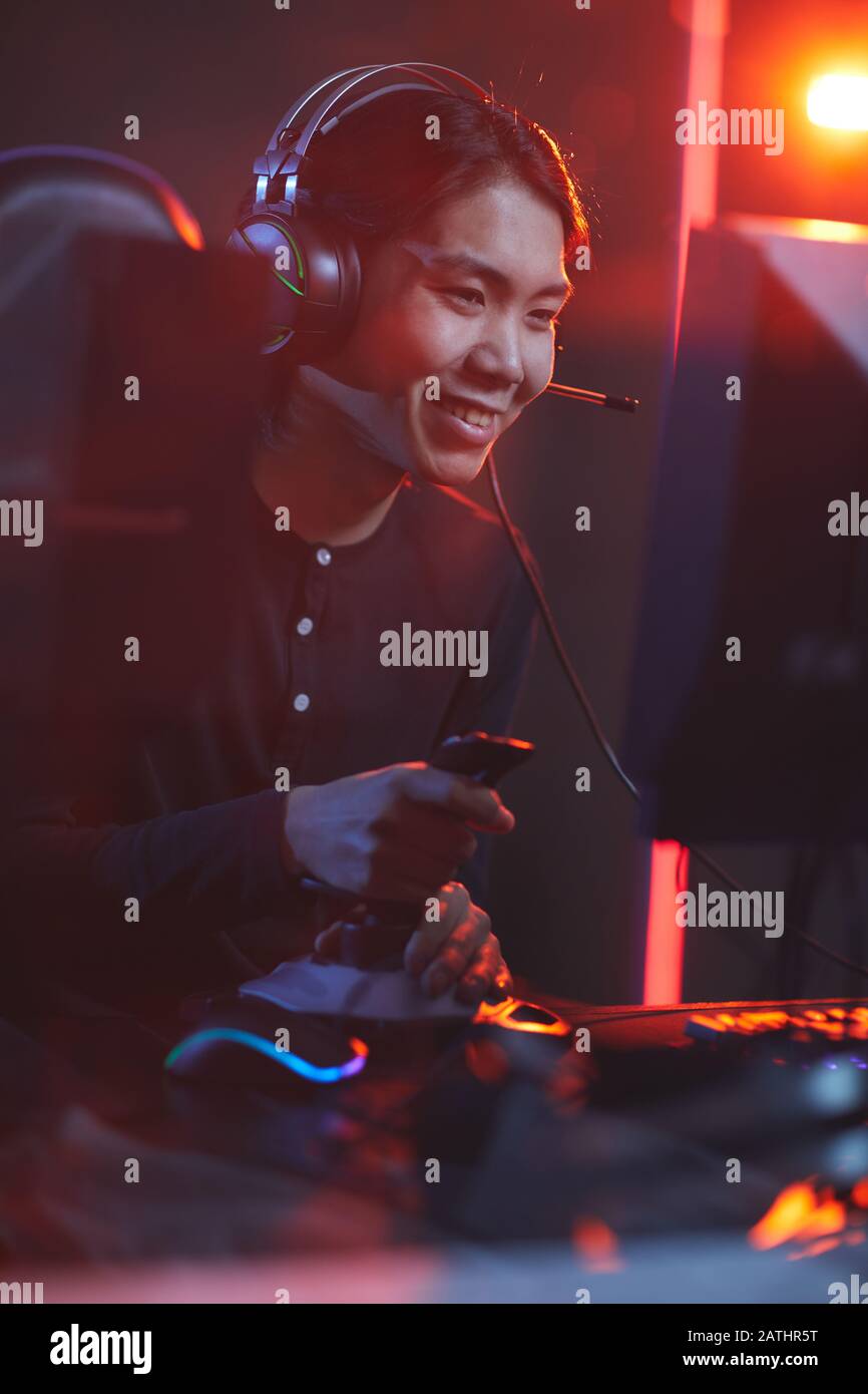 Ritratto verticale di uomo asiatico sorridente che gioca ai videogiochi utilizzando il cambio di corsa in interni scuri cyber Foto Stock