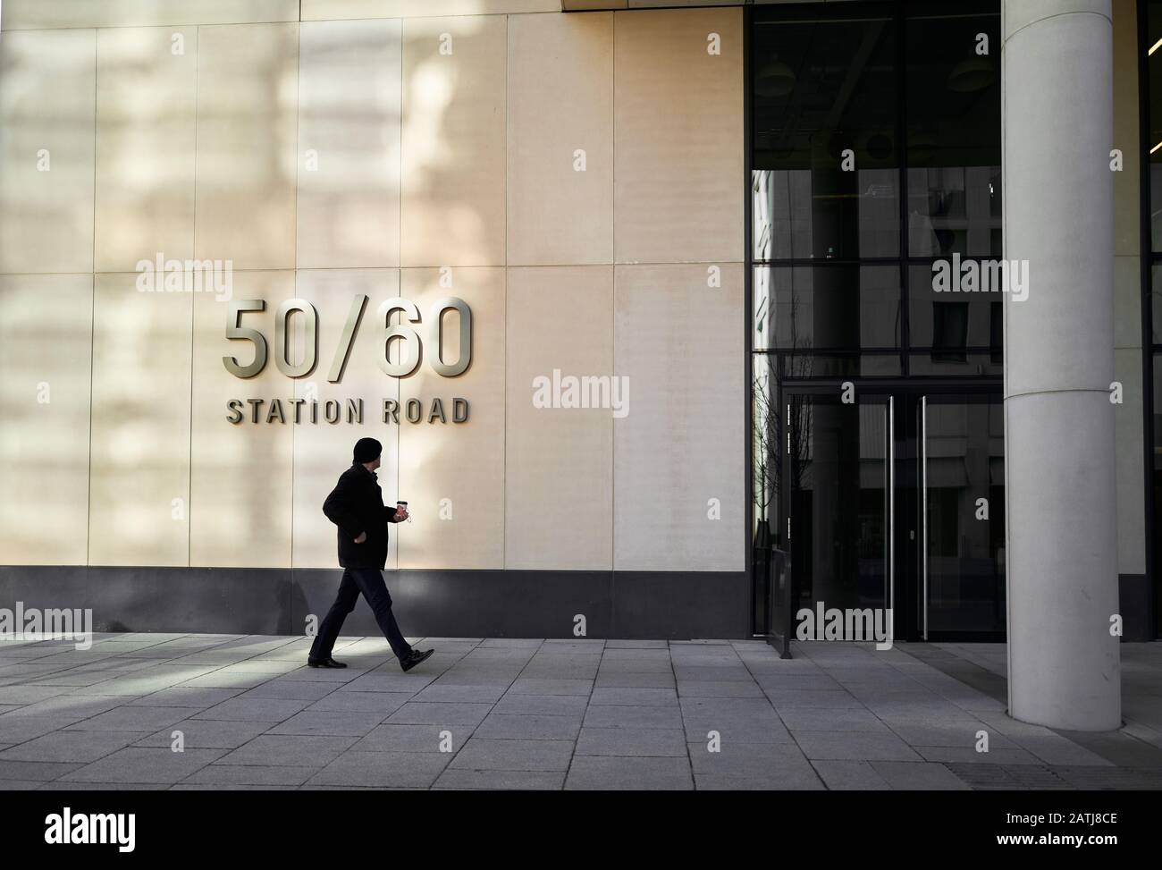 Un uomo che tiene una tazza di caffè passa la facciata illuminata dal sole del moderno blocco ufficio alla 50/60 Station Road, Cambridge, Inghilterra. Foto Stock