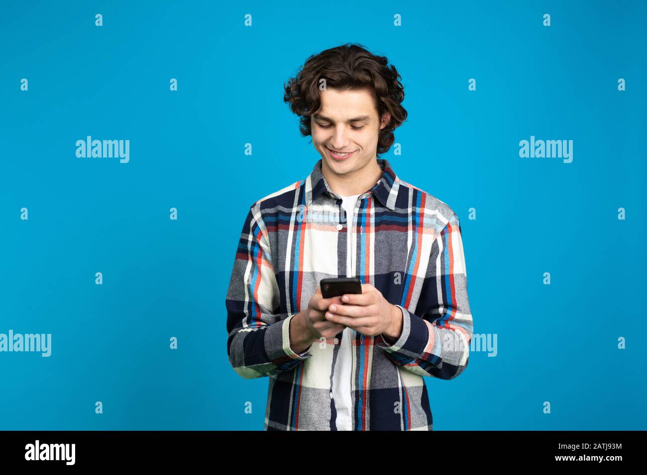 Sorridente ragazzo in camicia a scacchi utilizzando uno smartphone isolato su sfondo blu. Foto Stock Sorridente ragazzo in camicia a scacchi utilizzando uno smartphone isolato su sfondo blu. Foto Stock
