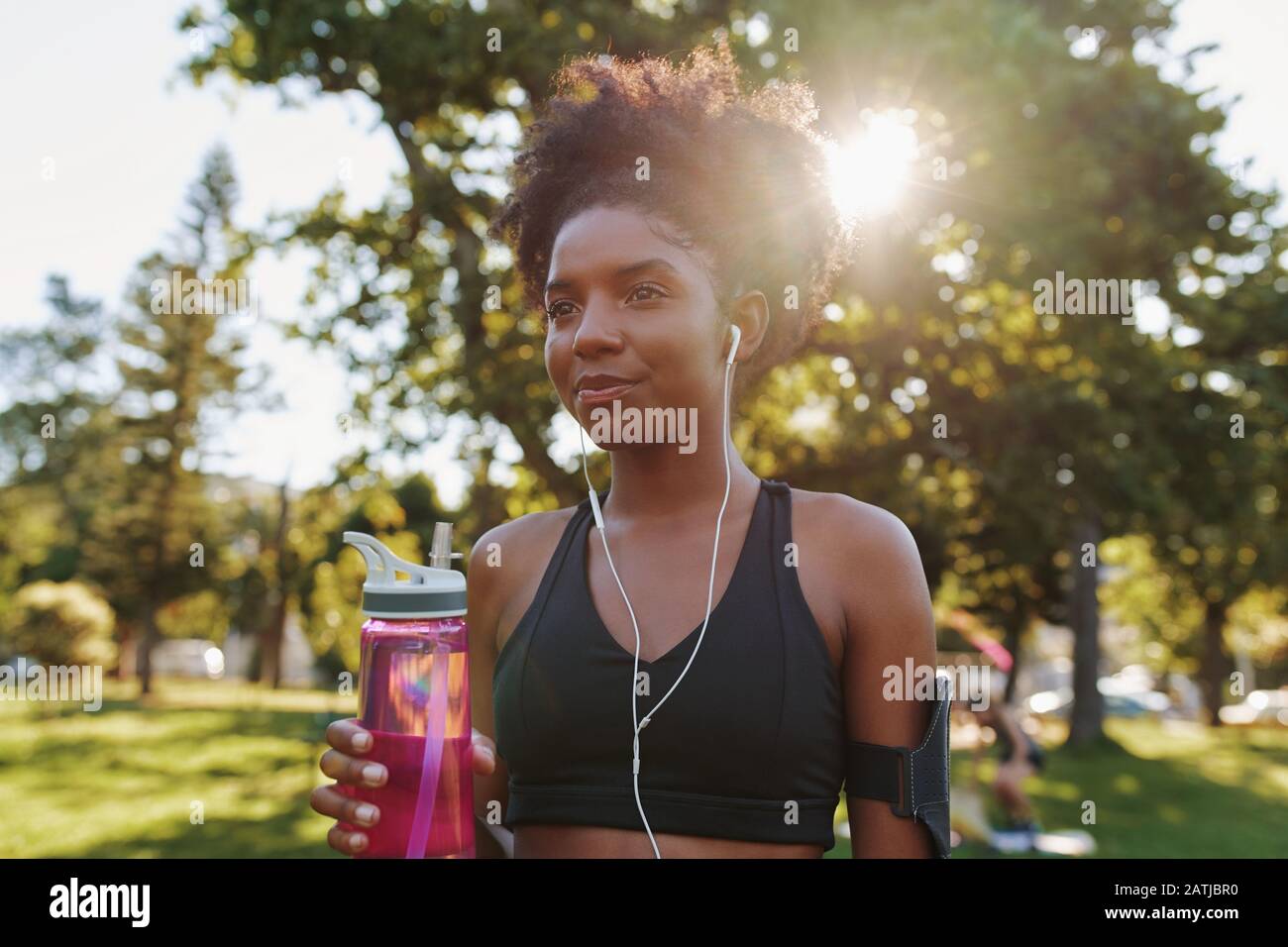 Atletica femminile in piedi in una luminosa giornata di sole nel parco con le cuffie nelle orecchie che tengono eco-friendly acqua potabile bottiglia in mano al parco Foto Stock