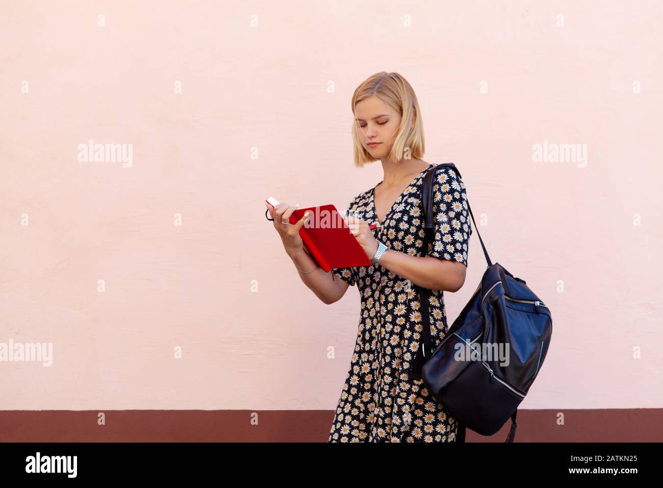 Ritratto di ragazza studentesca sorridente di moda con zaino nero e tenendo pianificatore rosso sul rosa Foto Stock