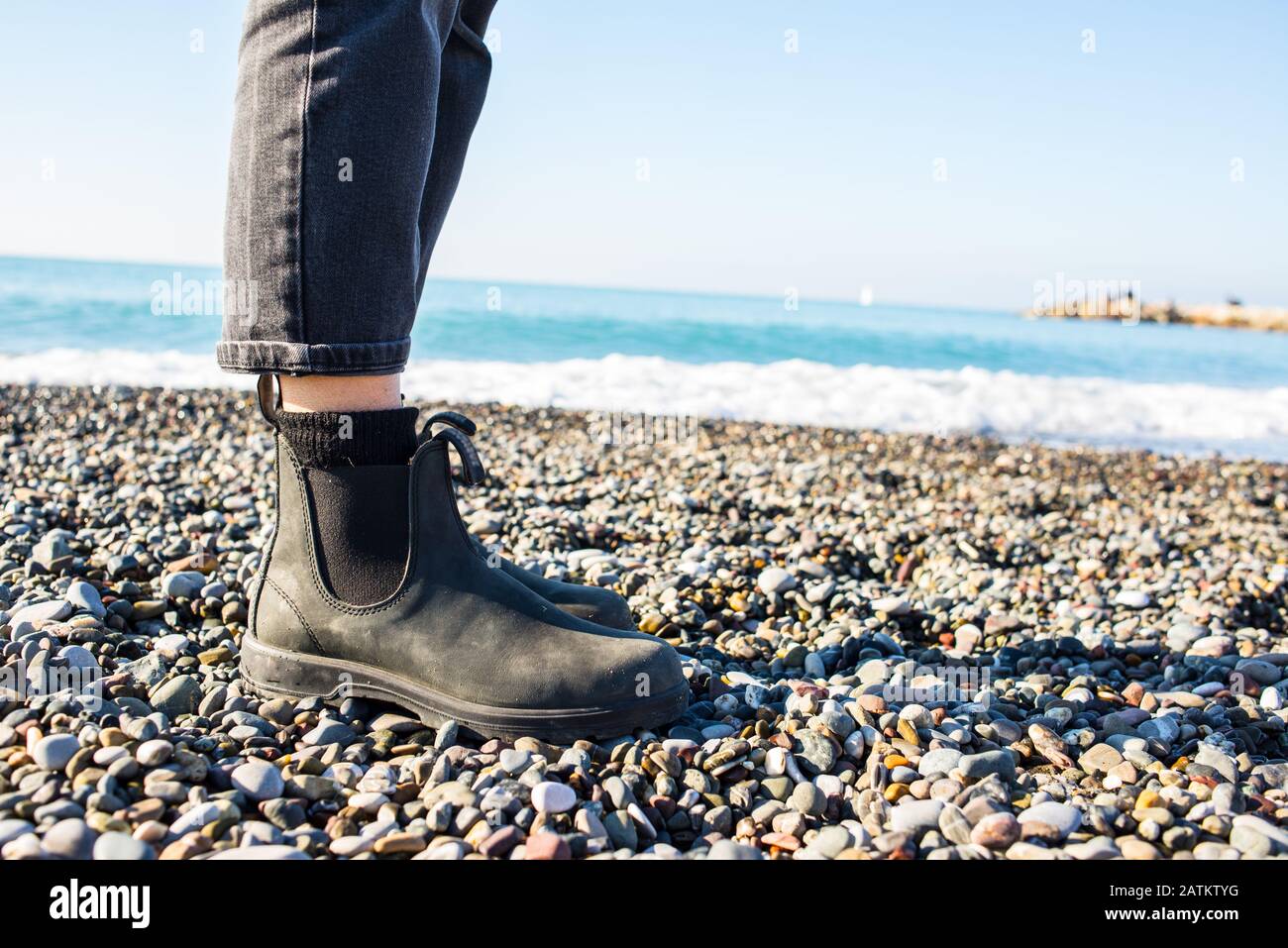 Stivali Chelsea classica suola in gomma nera. Concentrati sulle gambe di una donna hippster che indossa pantaloni neri denim. Sparato sulla spiaggia di ghiaia con il mare in grigro Foto Stock